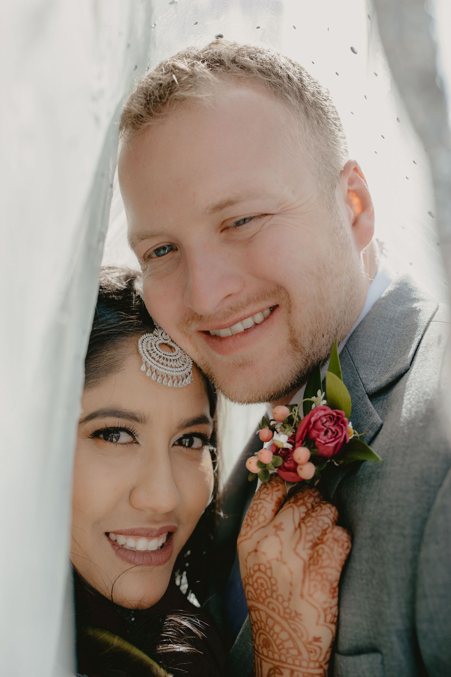 Bride with mehndi and chandelier earring poses with groom in gray suit and pink rose boutonniere at wedding.