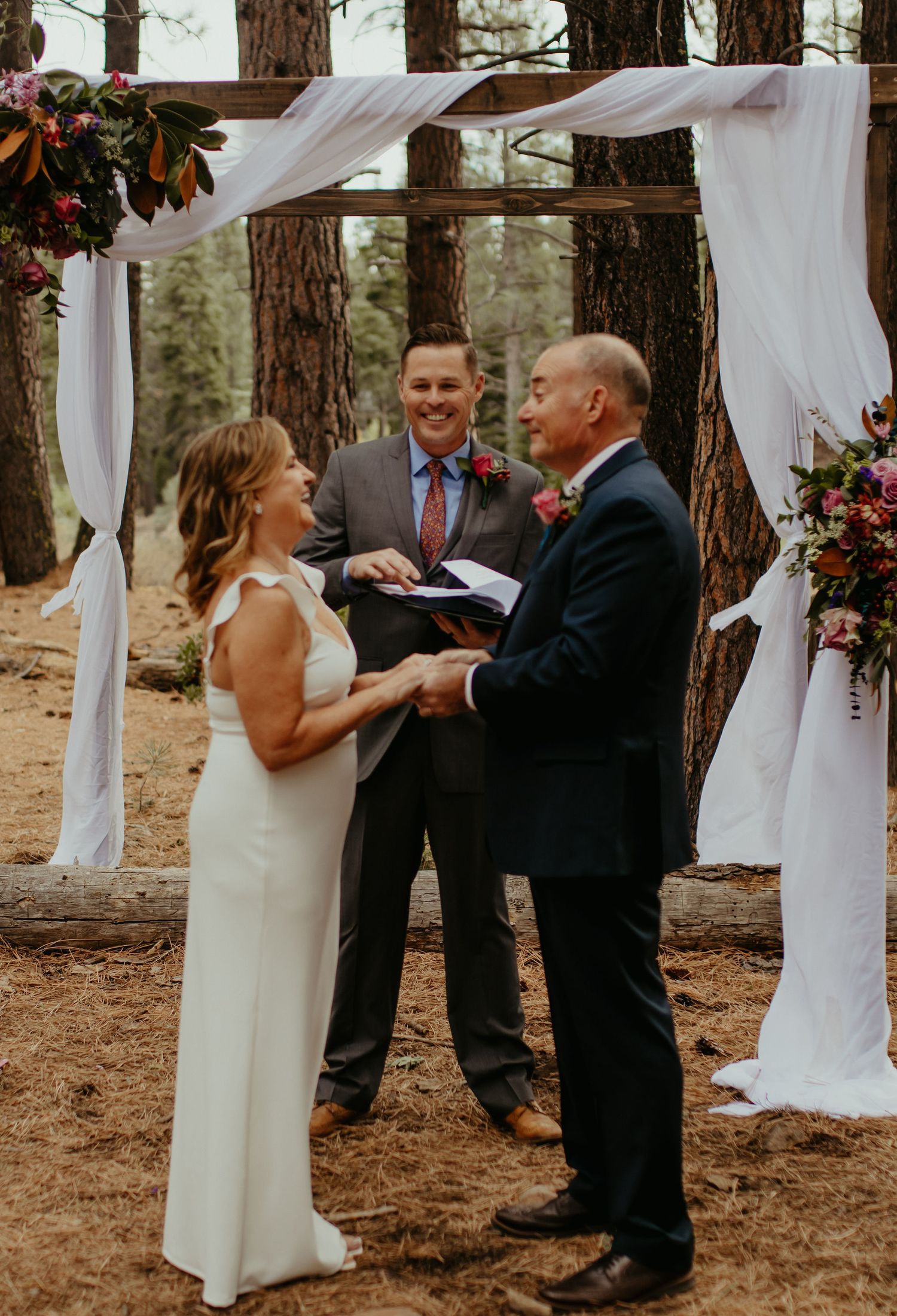 Outdoor wedding ceremony in a pine forest with bride and groom exchanging vows under a rustic wooden arch with white draping.