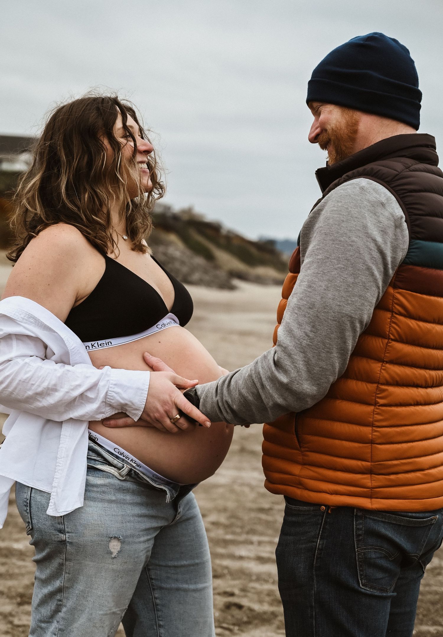 Expecting couple smiles at each other on a beach as man touches pregnant belly.