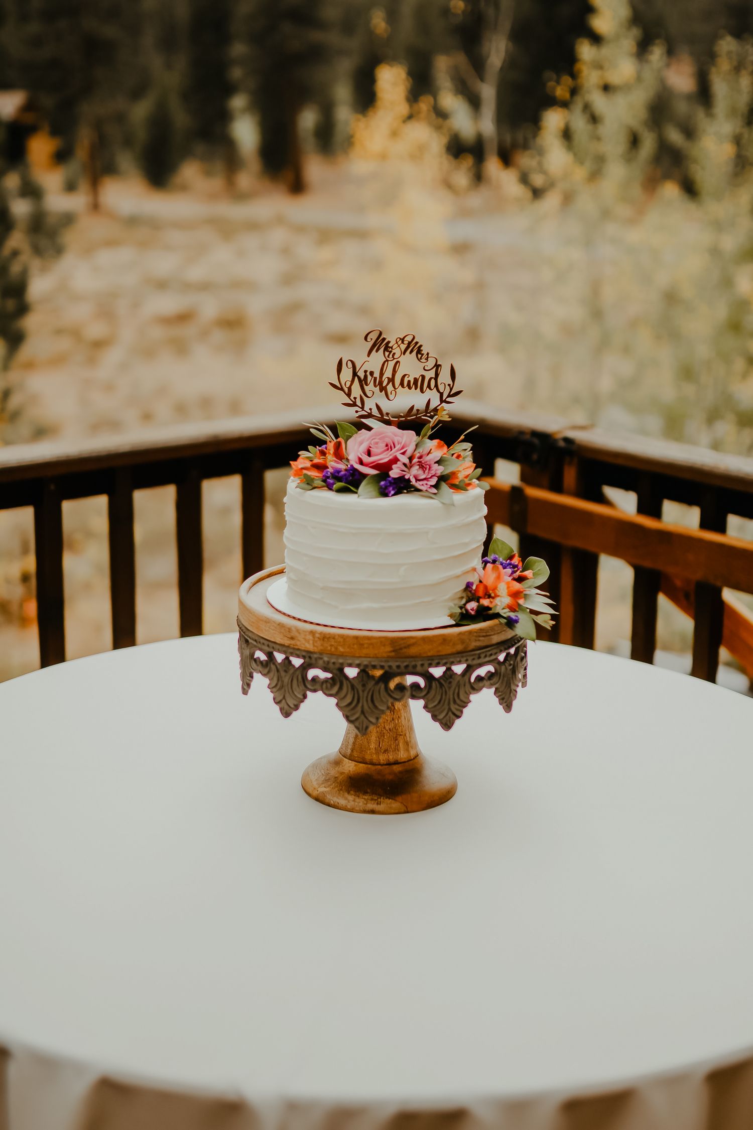 A single-tier white wedding cake with colorful floral topper on a gold cake stand displayed on a white table outdoors.