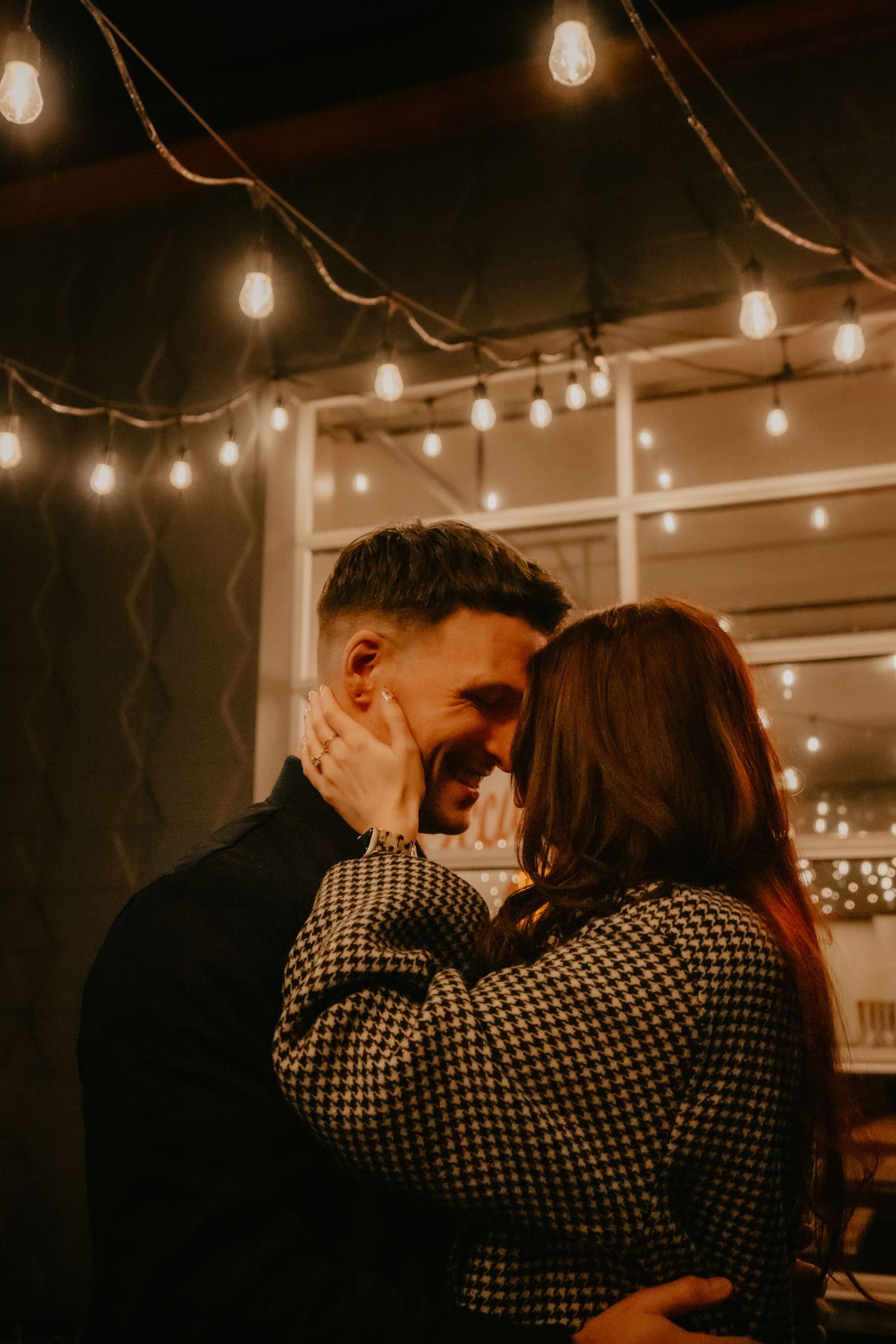 Romantic couple embracing and smiling under warm string lights in a cozy evening setting.