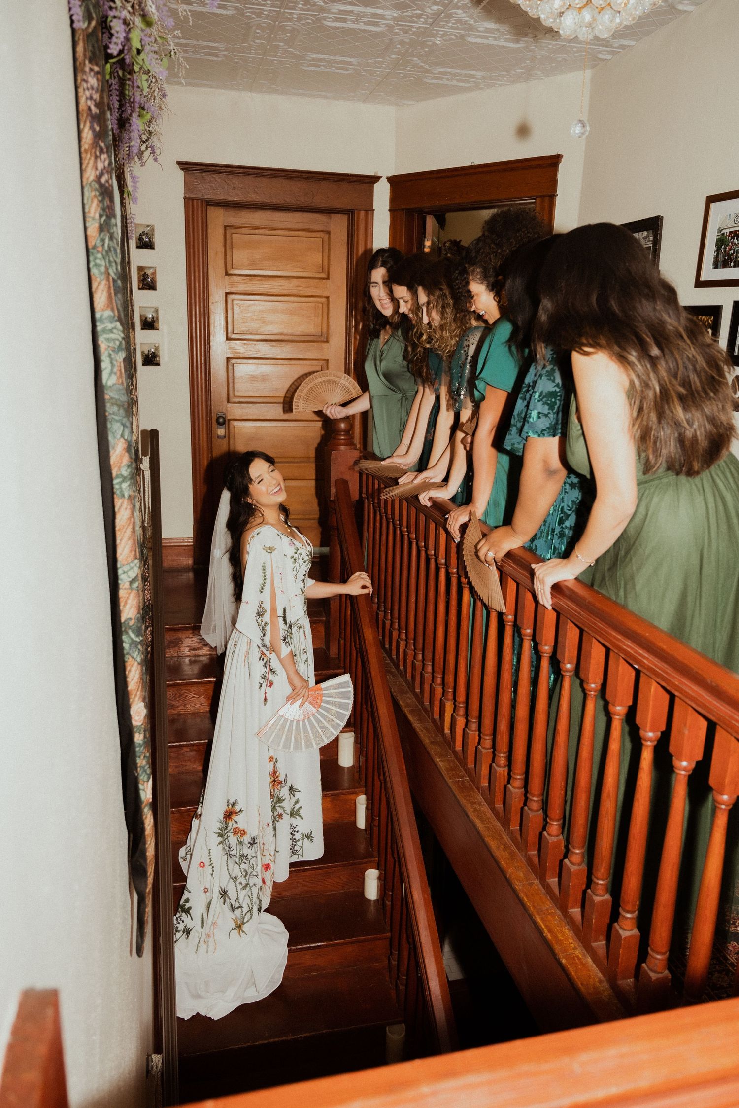 Bride in floral gown holds fan while bridesmaids in green dresses look down from wooden staircase railing.