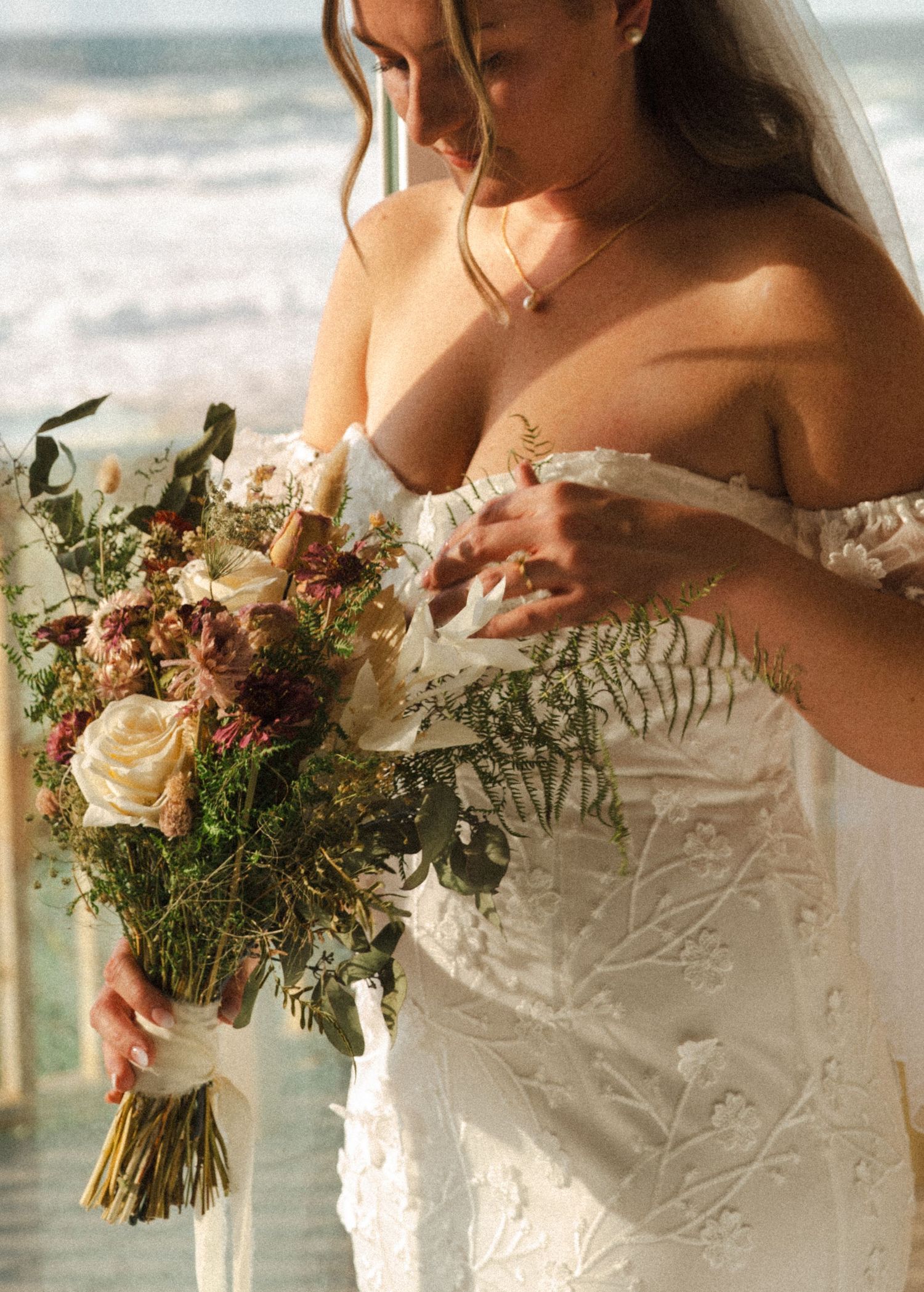 Bride in an off-the-shoulder lace wedding gown holds a rustic bouquet of roses and greenery at the beach.