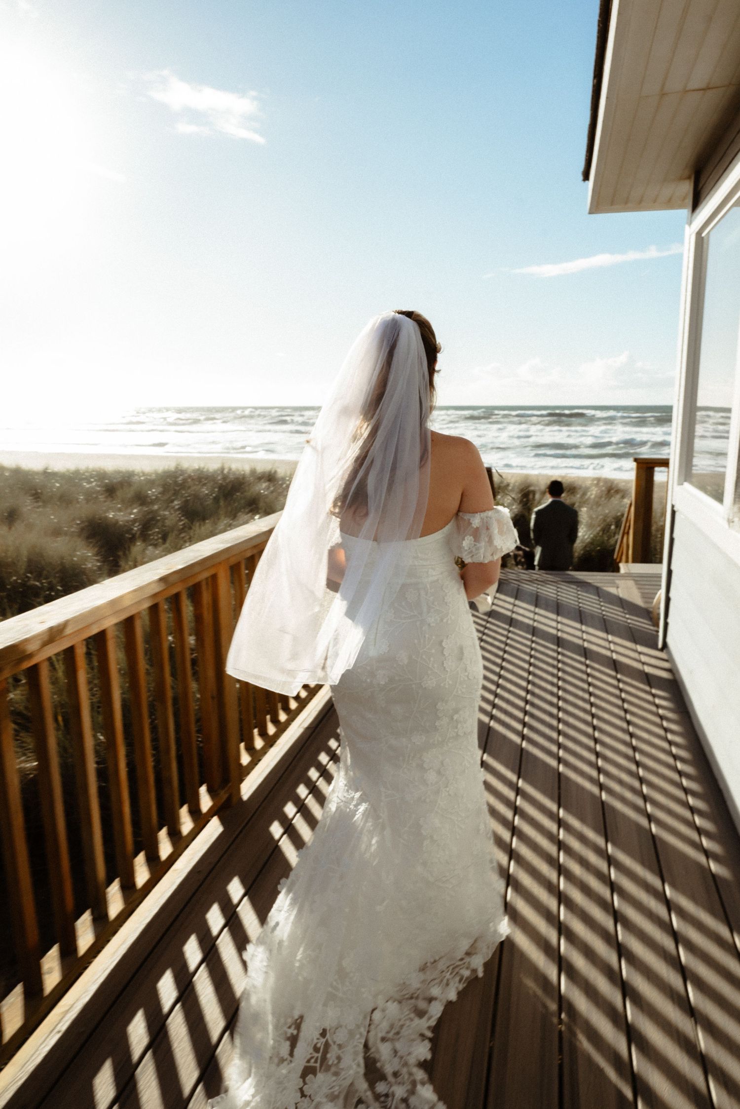 Bride in a lace mermaid gown and veil walks along a sunlit wooden beach deck toward the ocean on her wedding day.