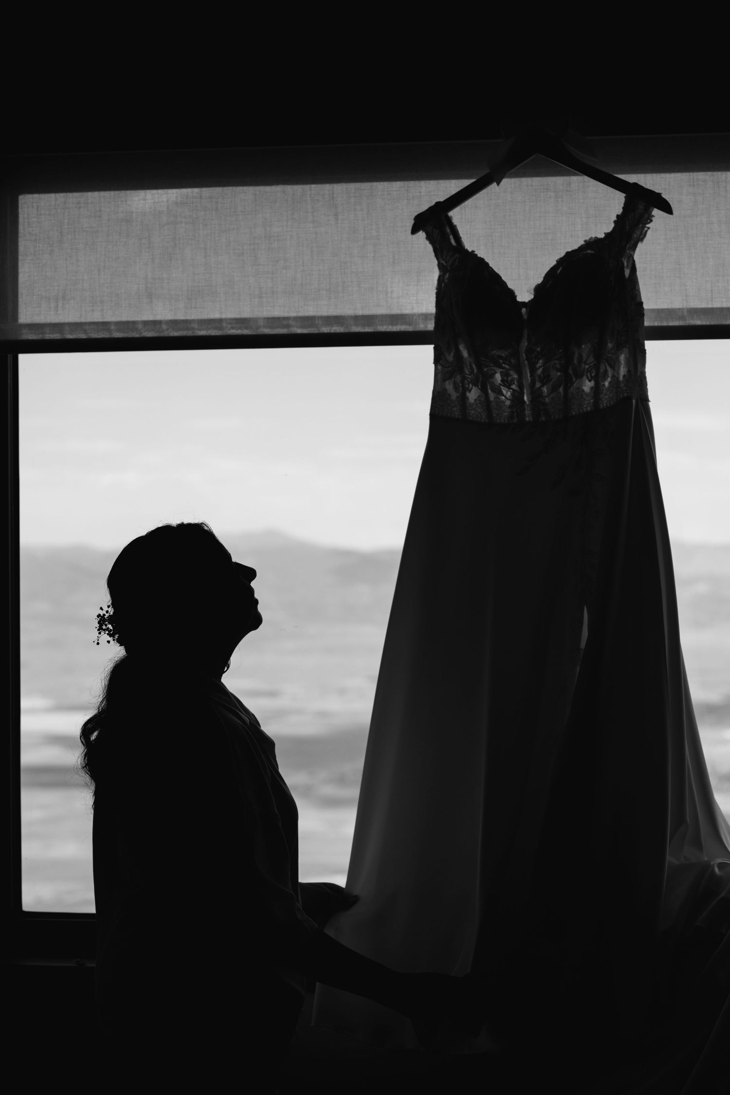 Black and white silhouette of a bride gazing at her lace wedding dress hanging by a window.