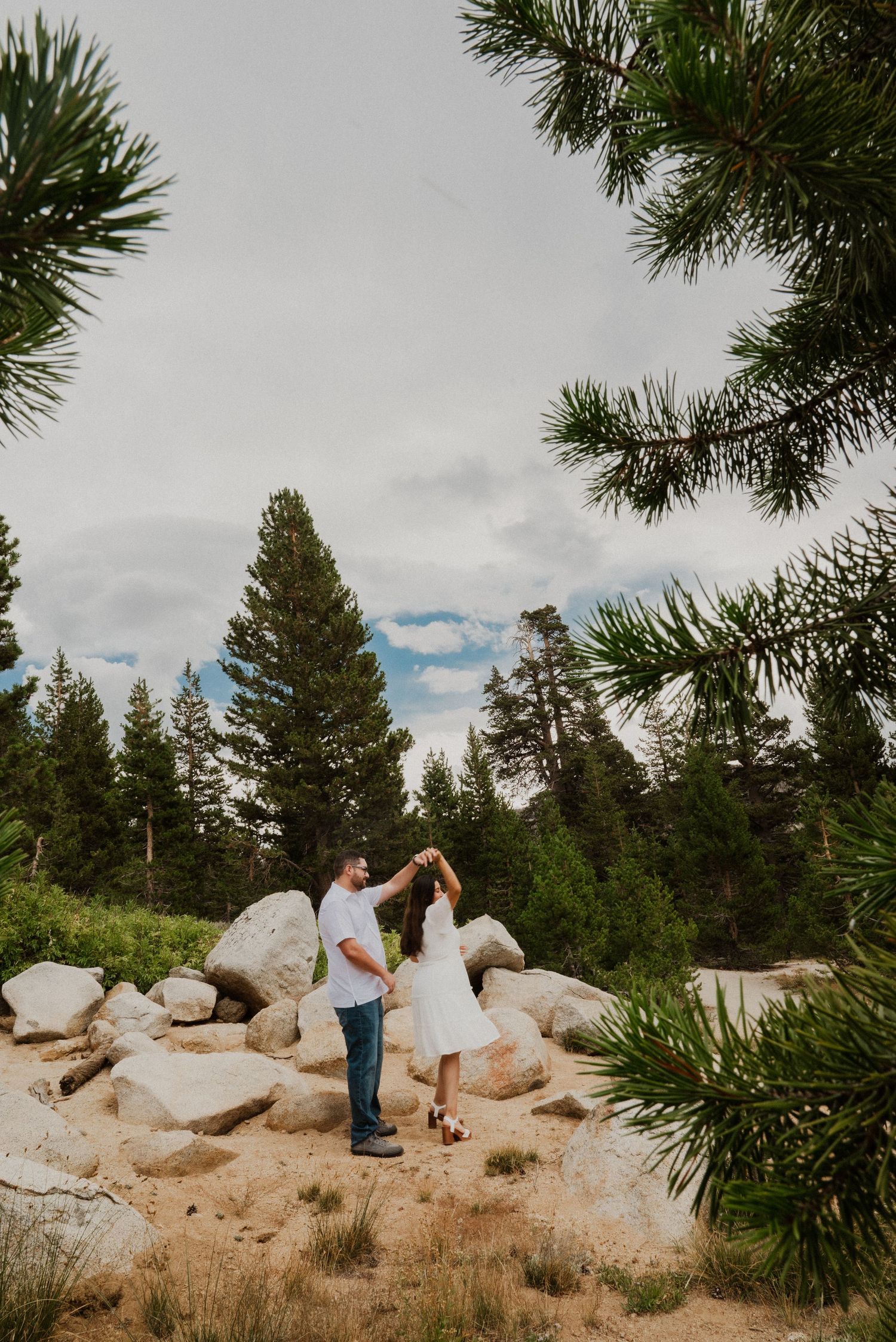 Couple dancing among boulders and pine trees in a scenic forest setting, woman in white dress, man in jeans.