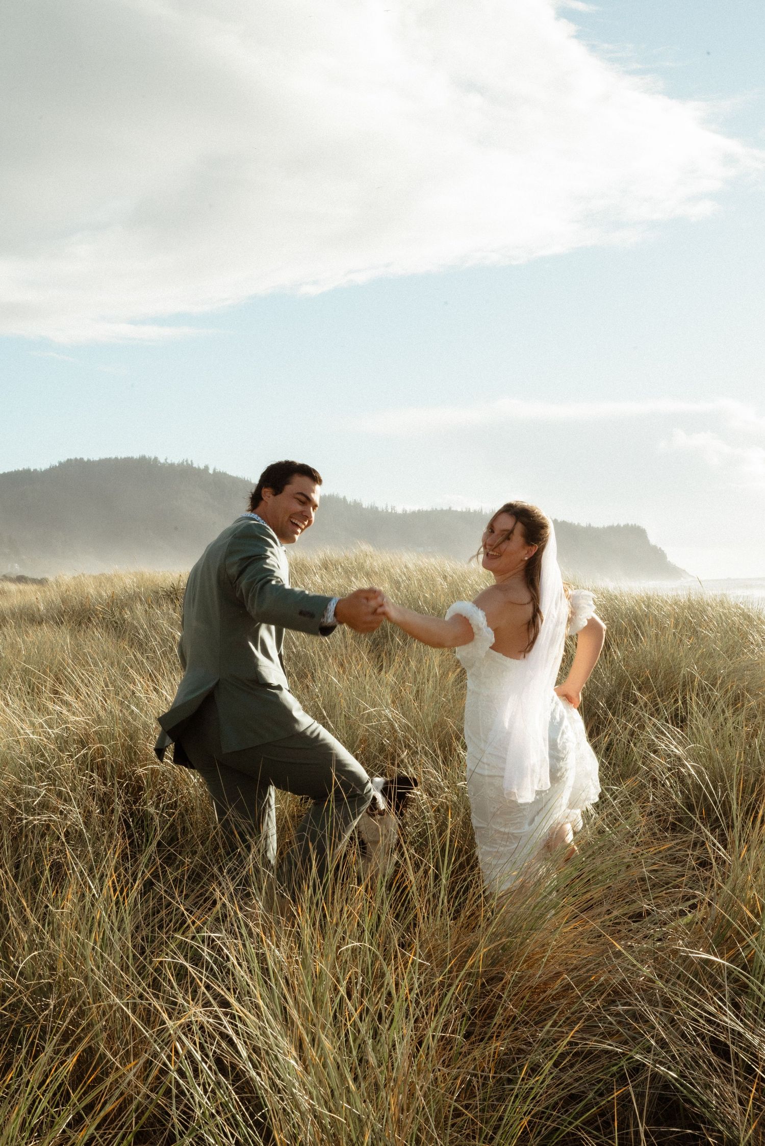 A smiling groom in a green suit leads his bride in a white gown by the hand through golden beach grass on a sunny coastal day.