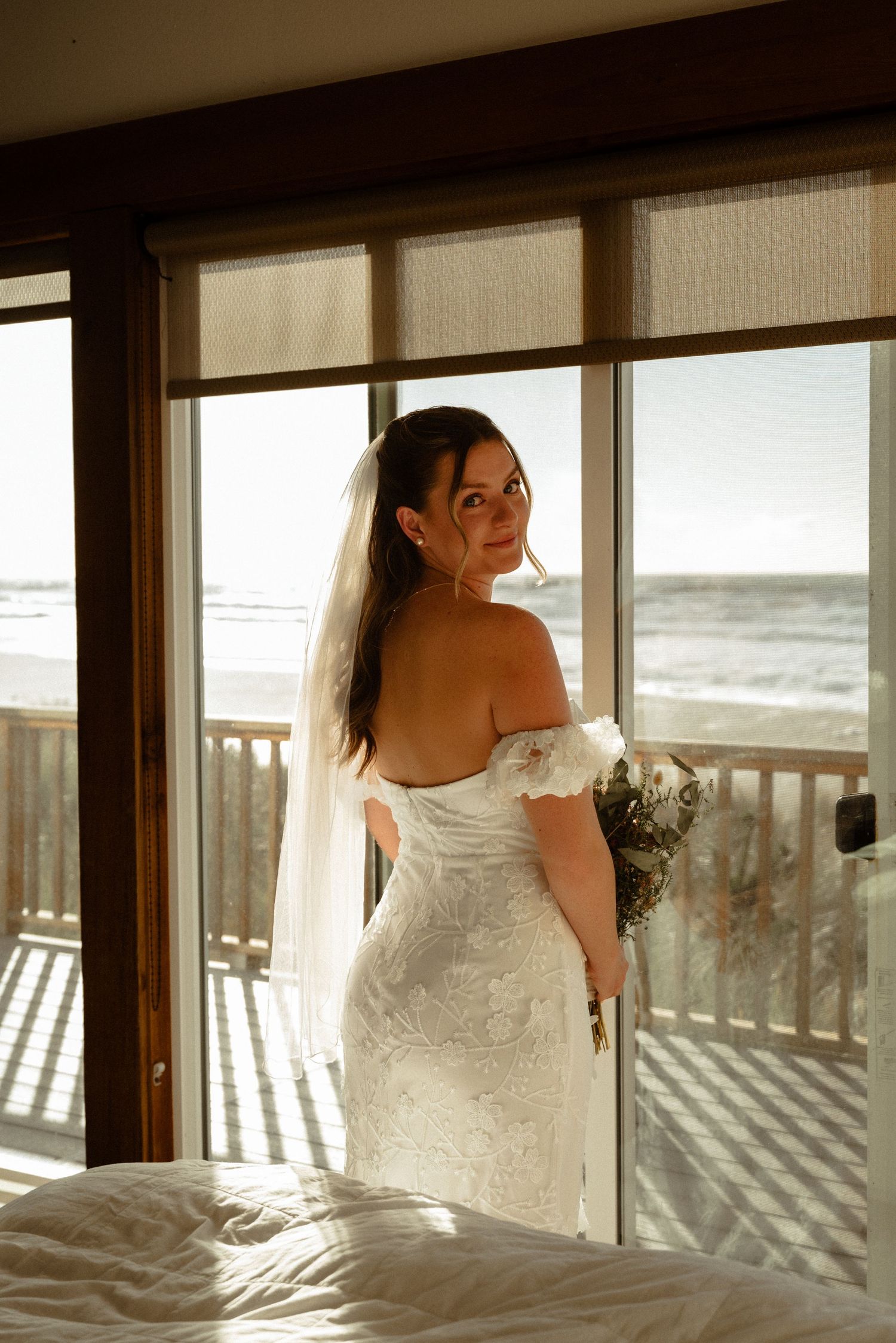 Bride in a lace off-the-shoulder wedding gown and long veil glances back, holding a bouquet by oceanview windows.