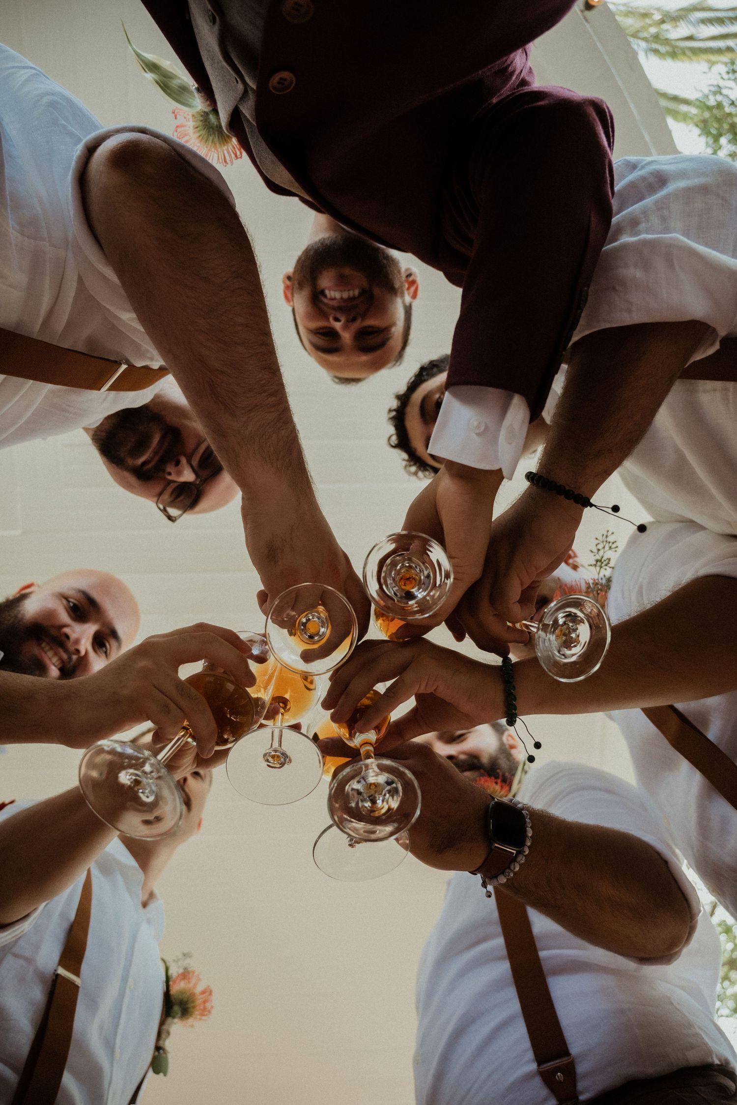 Groomsmen in white shirts toast with wine glasses in a circle, viewed from below at a wedding celebration.