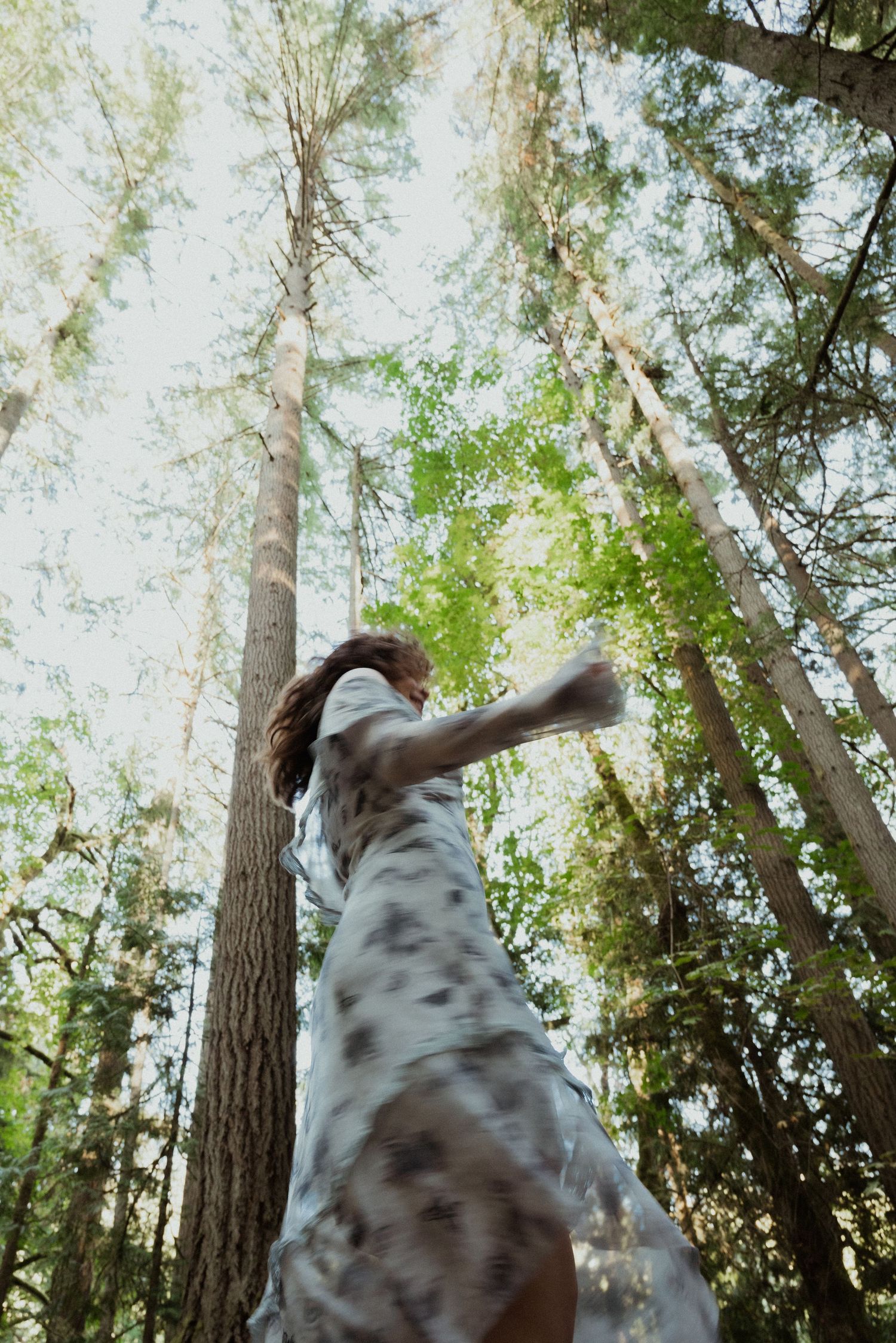 Woman in spotted dress twirling among tall trees in a forest, shot from low angle looking up.