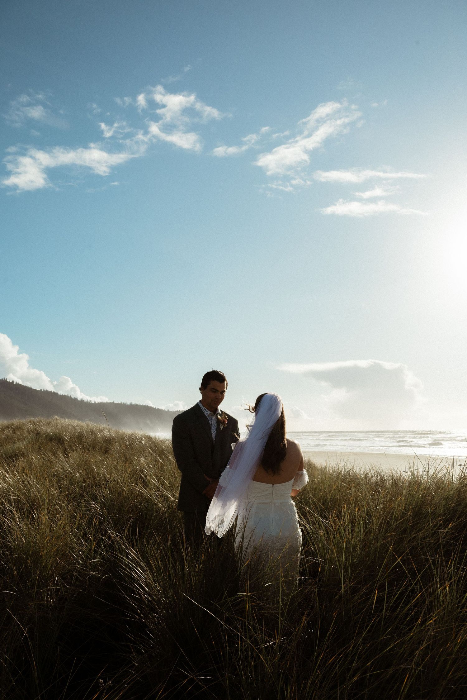 Silhouetted bride and groom stand together in beach dune grass as the sun glows over the ocean.