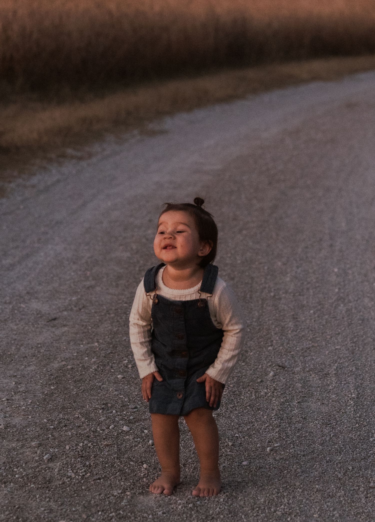 Adorable toddler girl in overalls standing barefoot on a gravel road at golden hour.