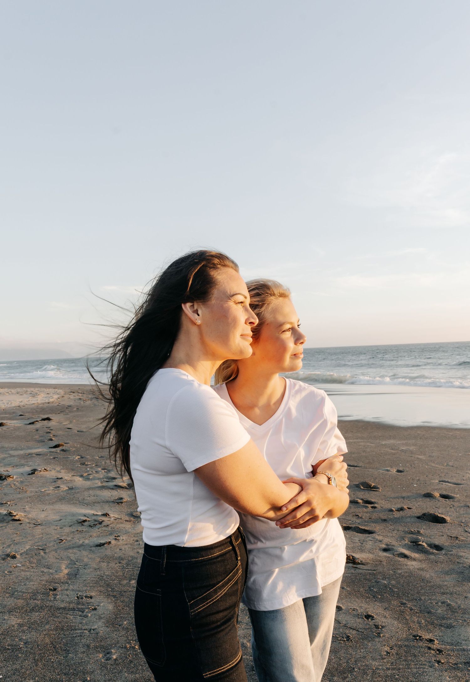 Two women in white t-shirts embrace on a beach at sunset, hair blowing in the wind with ocean waves behind them.