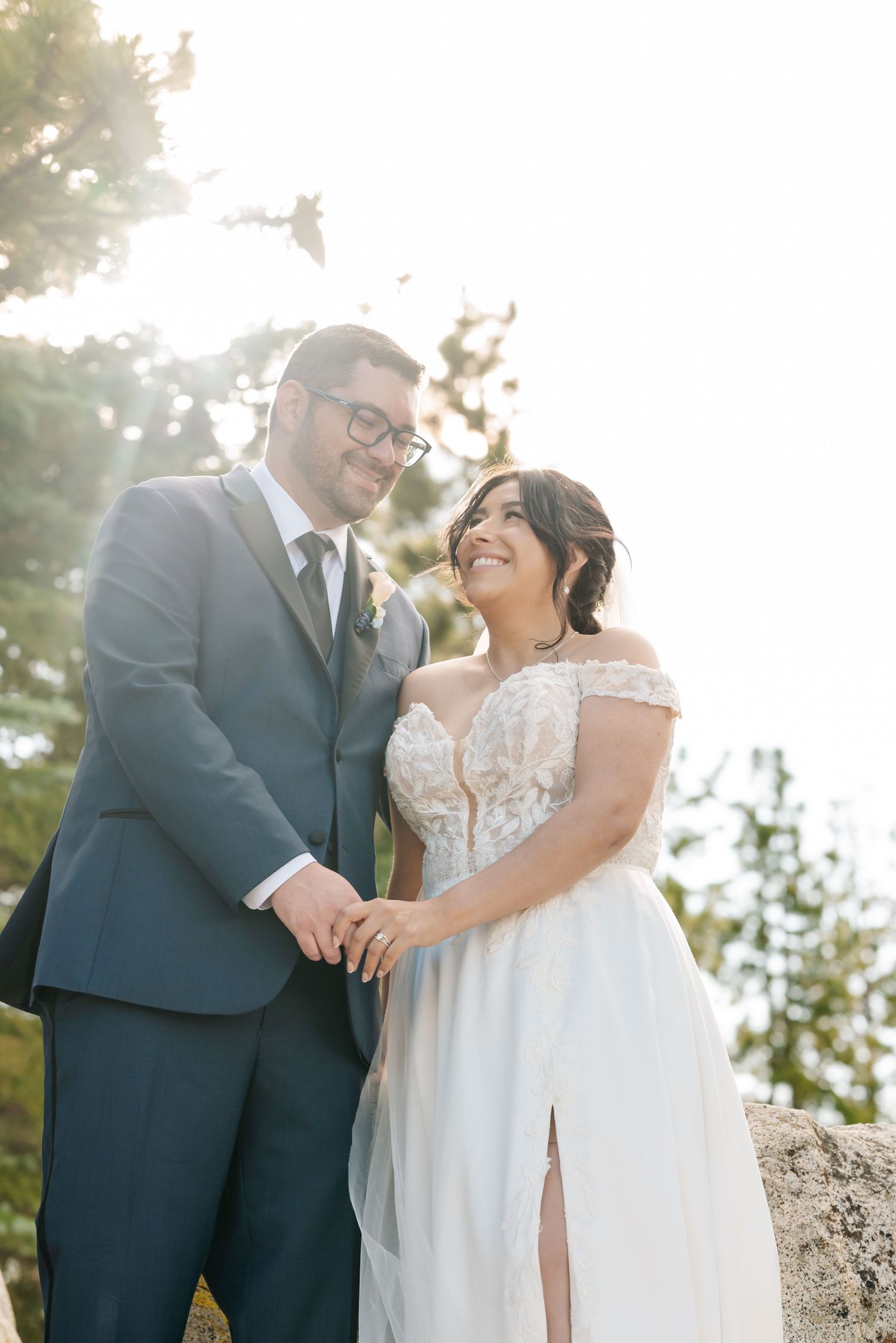 A smiling bride in a lace off-shoulder gown holds hands with a groom in a navy suit outdoors in golden sunlight.