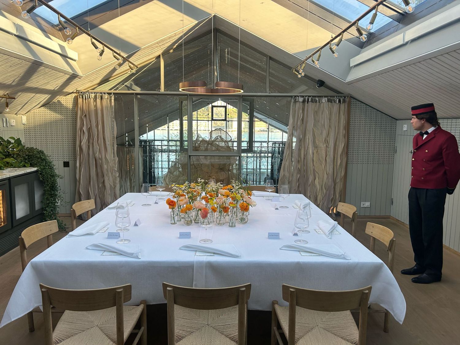 White-tablecloth dining table with floral arrangements in a glass conservatory with a uniformed staff member.