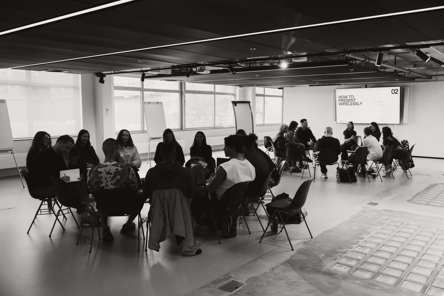 Black and white photo of a group workshop with participants seated at round tables in a modern open space.