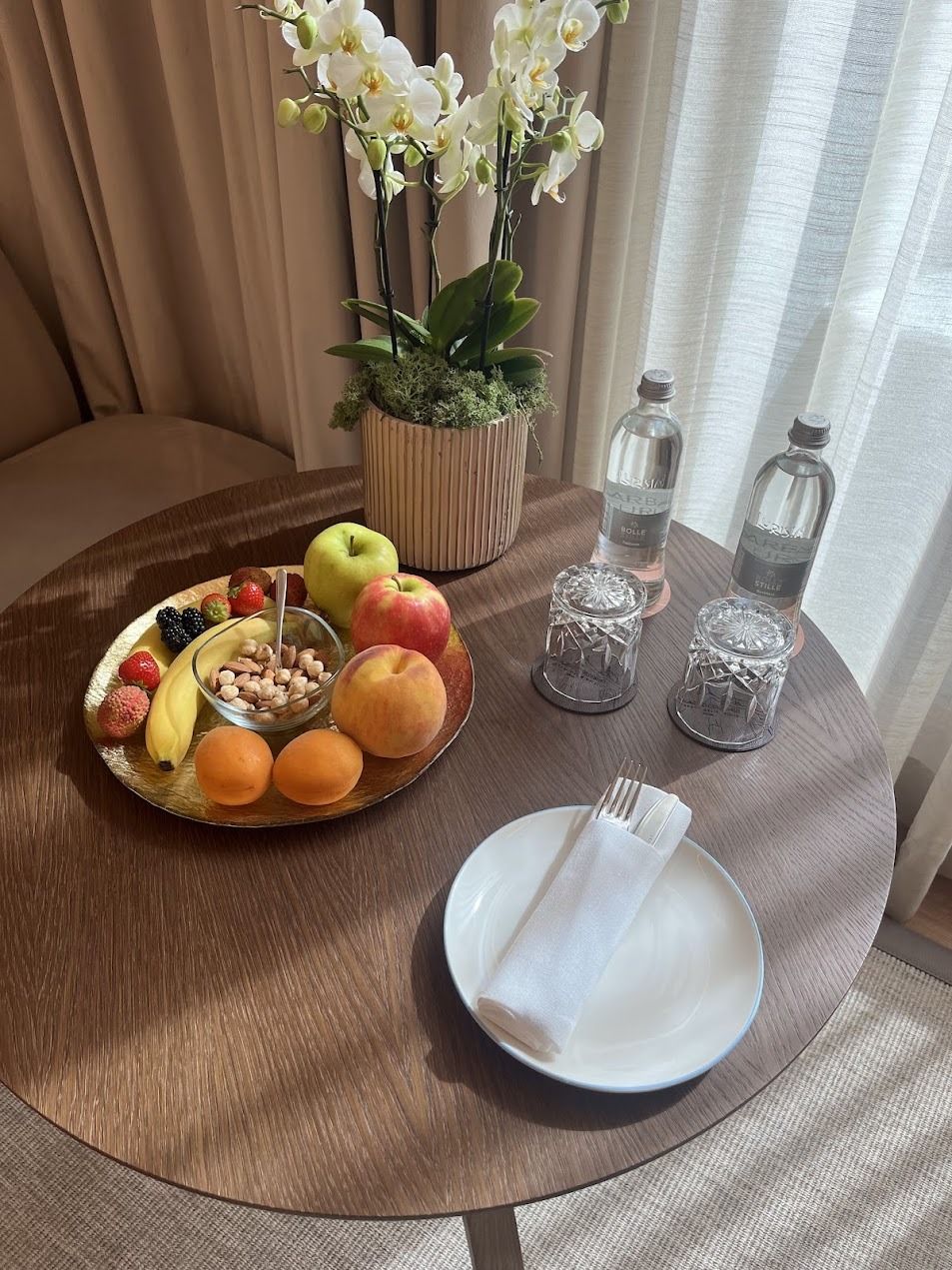 Hotel room table with fresh fruit platter, white orchids, water bottles, and place setting.
