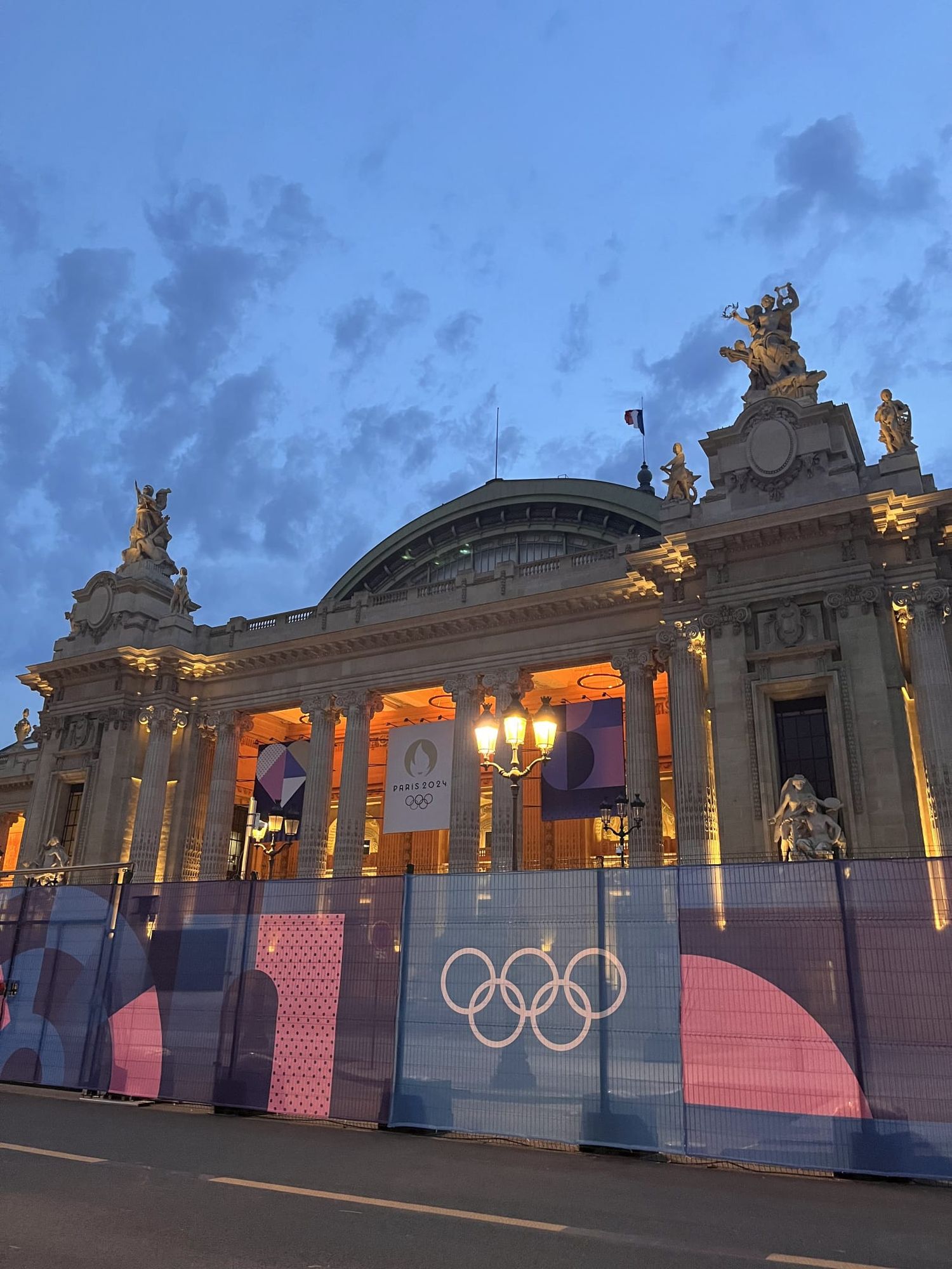 Grand Palais Paris illuminated at dusk with Olympic rings on construction barriers.