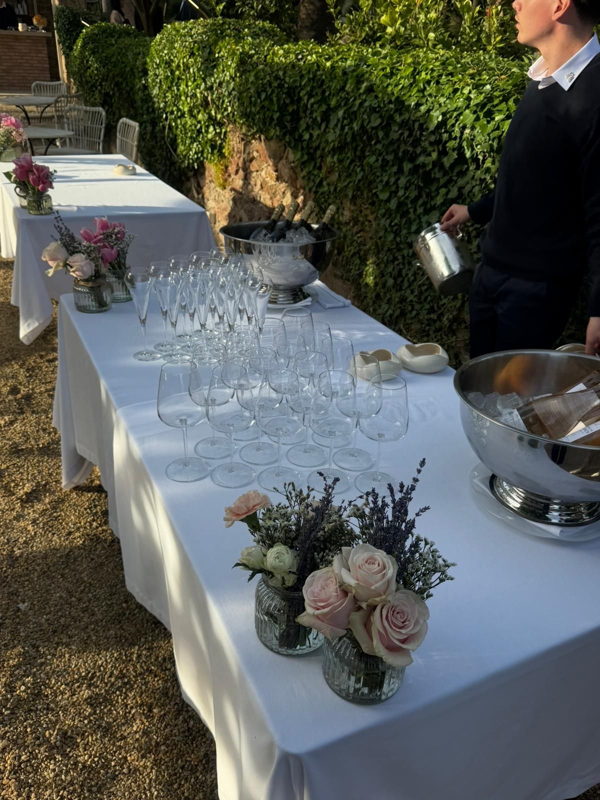 Outdoor bar setup with crystal wine glasses and pink rose floral arrangements on white tablecloth.