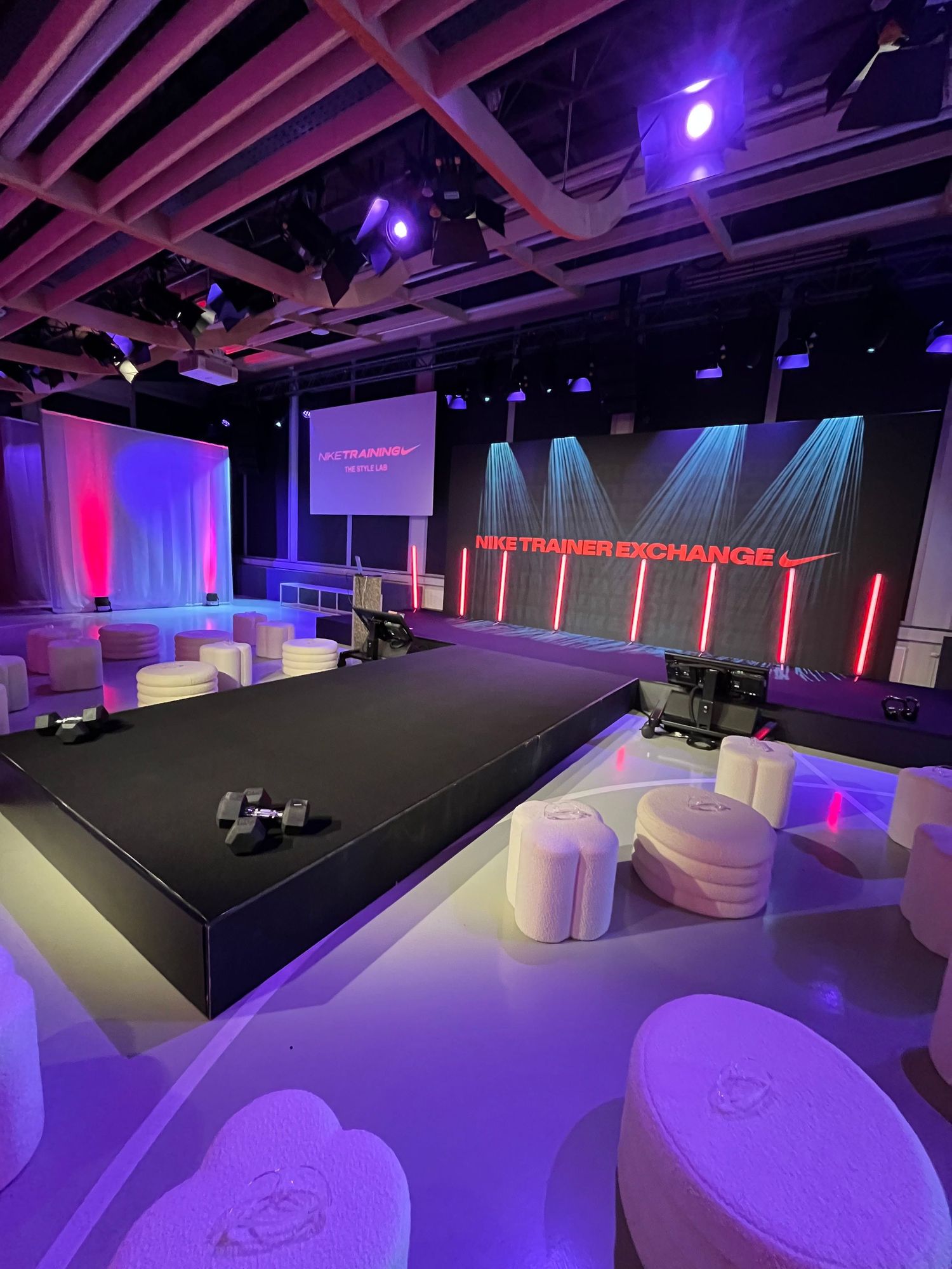 Modern fitness studio stage setup with white ottomans, dumbbells, and Nike Trainer Exchange signage under purple lighting.
