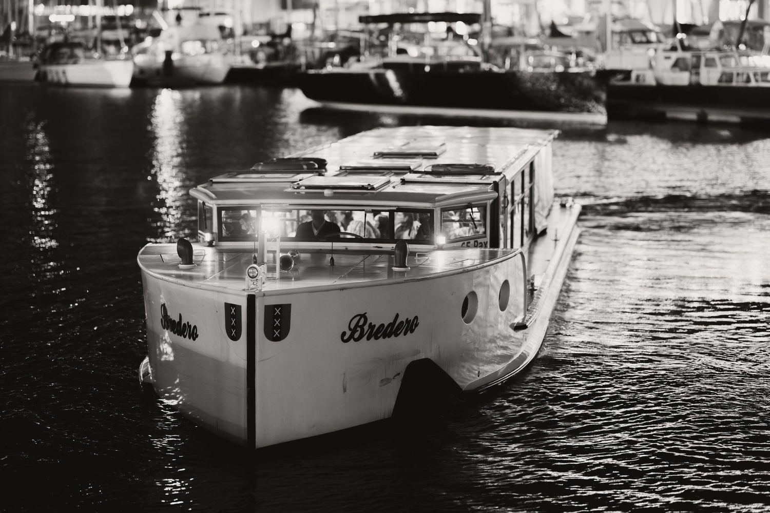 Black and white photo of illuminated Bredero boat navigating harbor at night with yachts in background.