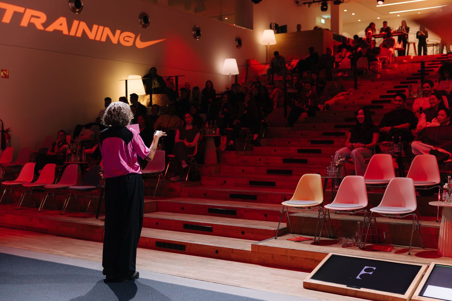 Speaker presenting to an audience at a Nike Training event with red ambient lighting and colorful chairs.
