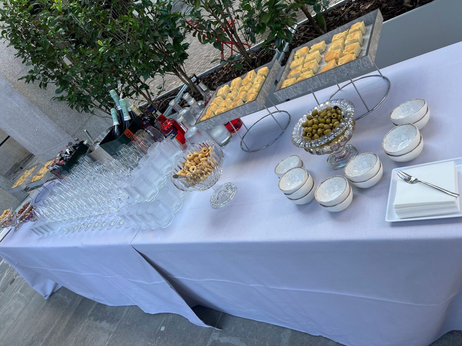 Outdoor catering table with champagne flutes, olives, popcorn, pastries, and desserts on white linen.