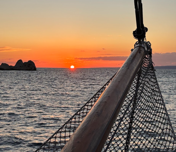 Golden sunset over calm ocean waters viewed from a sailboat bow with rope netting detail.