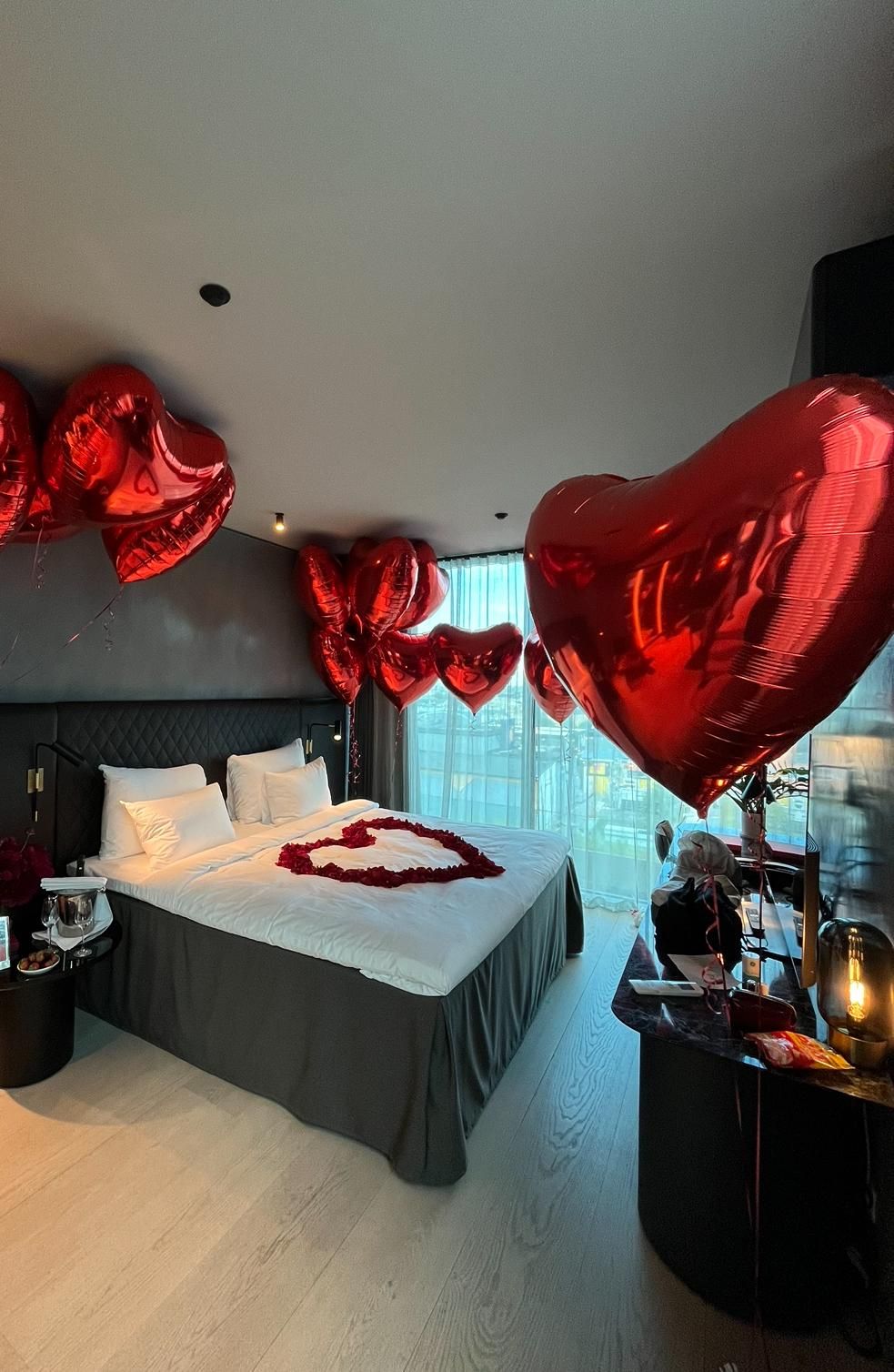 Romantic hotel room decorated with red heart balloons and rose petals on white bed.