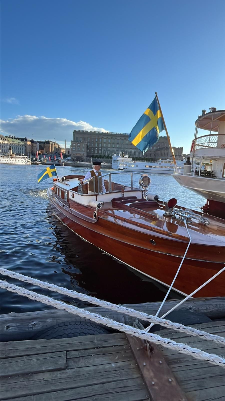 Classic wooden boat with Swedish flag docked at sunny Stockholm waterfront on clear blue day.