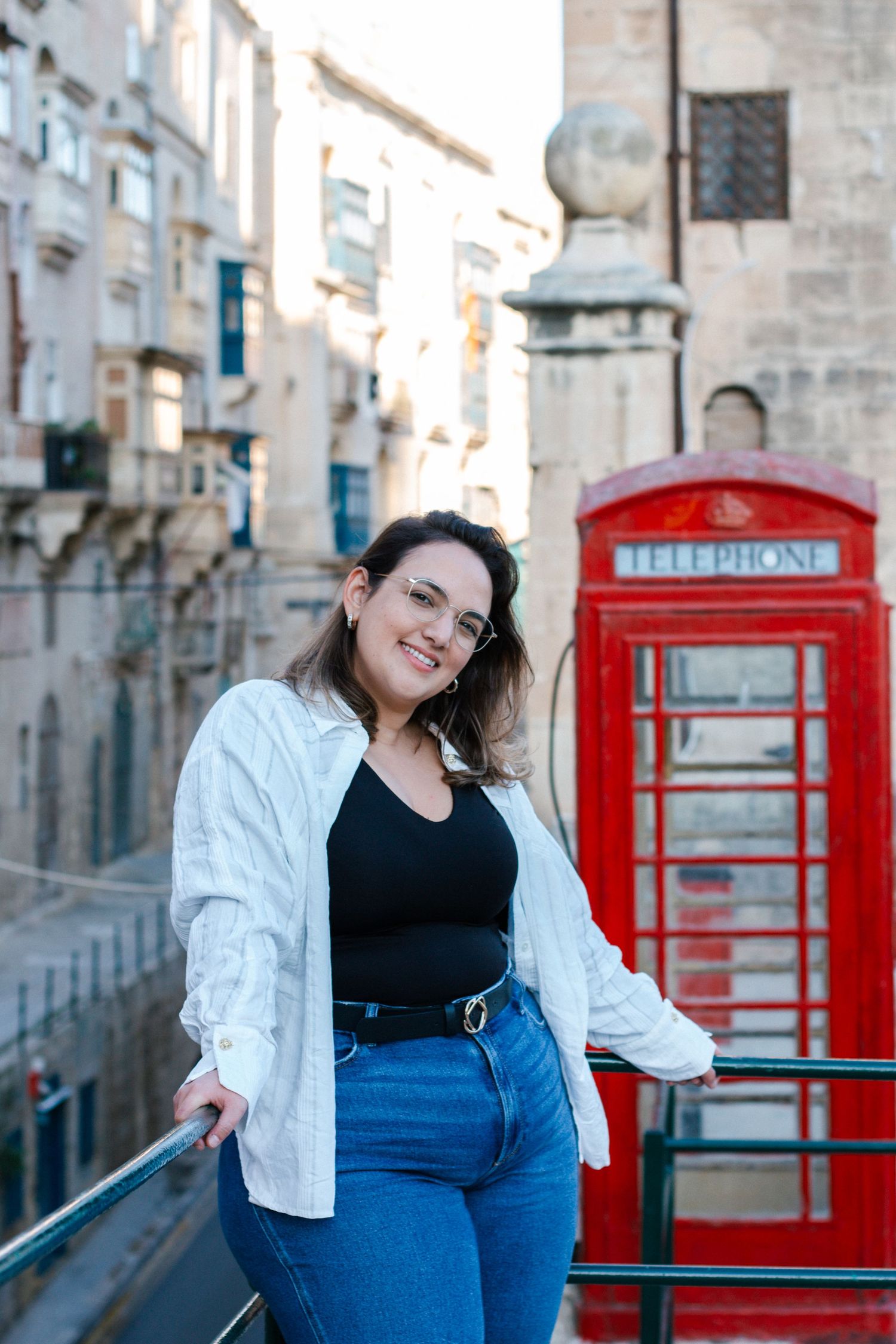 Smiling woman in white shirt leaning on railing with iconic red British telephone box behind her.