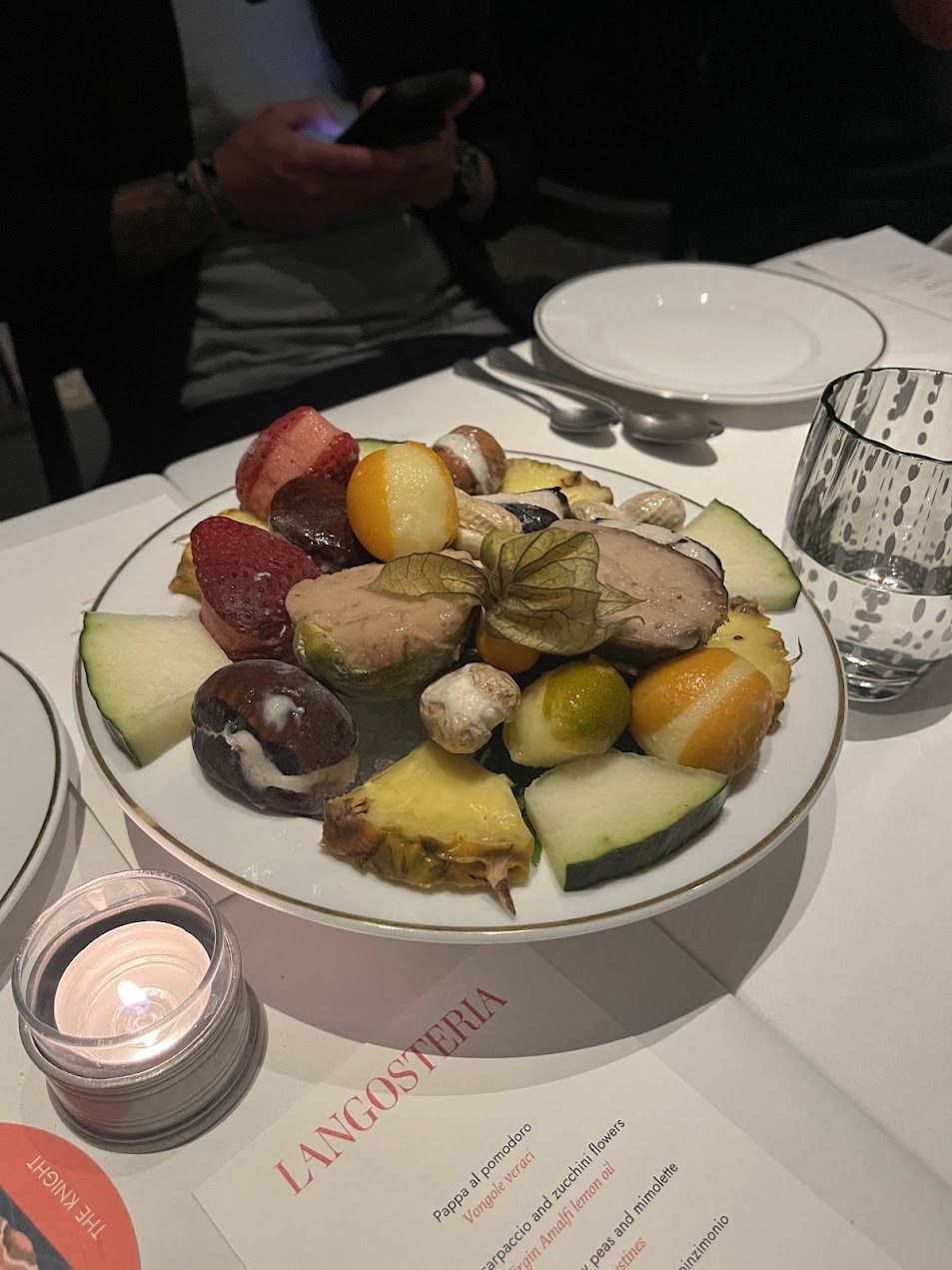 Elegant fruit and appetizer plate at Langosteria restaurant with candle and menu on white tablecloth.