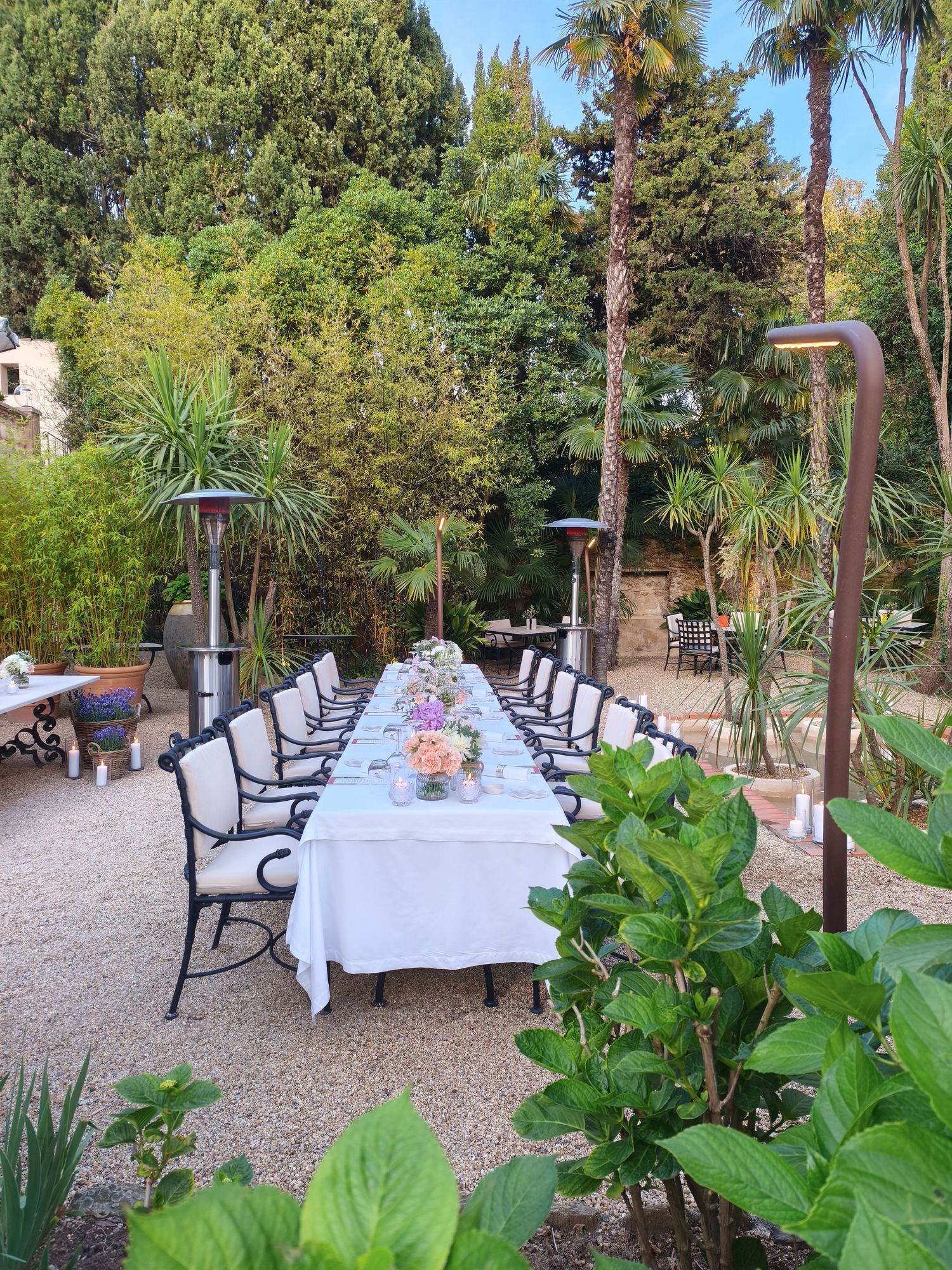 Long outdoor dining table set with white linens surrounded by lush garden greenery.