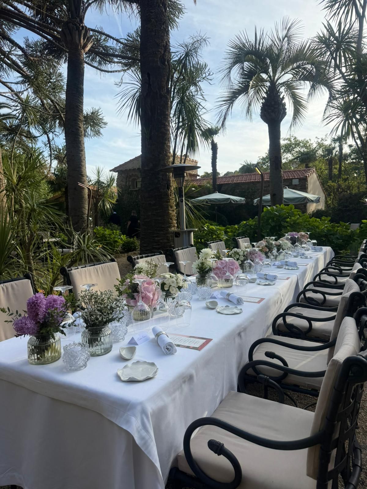 Elegant outdoor dining table set with white linens and floral centerpieces surrounded by palm trees.