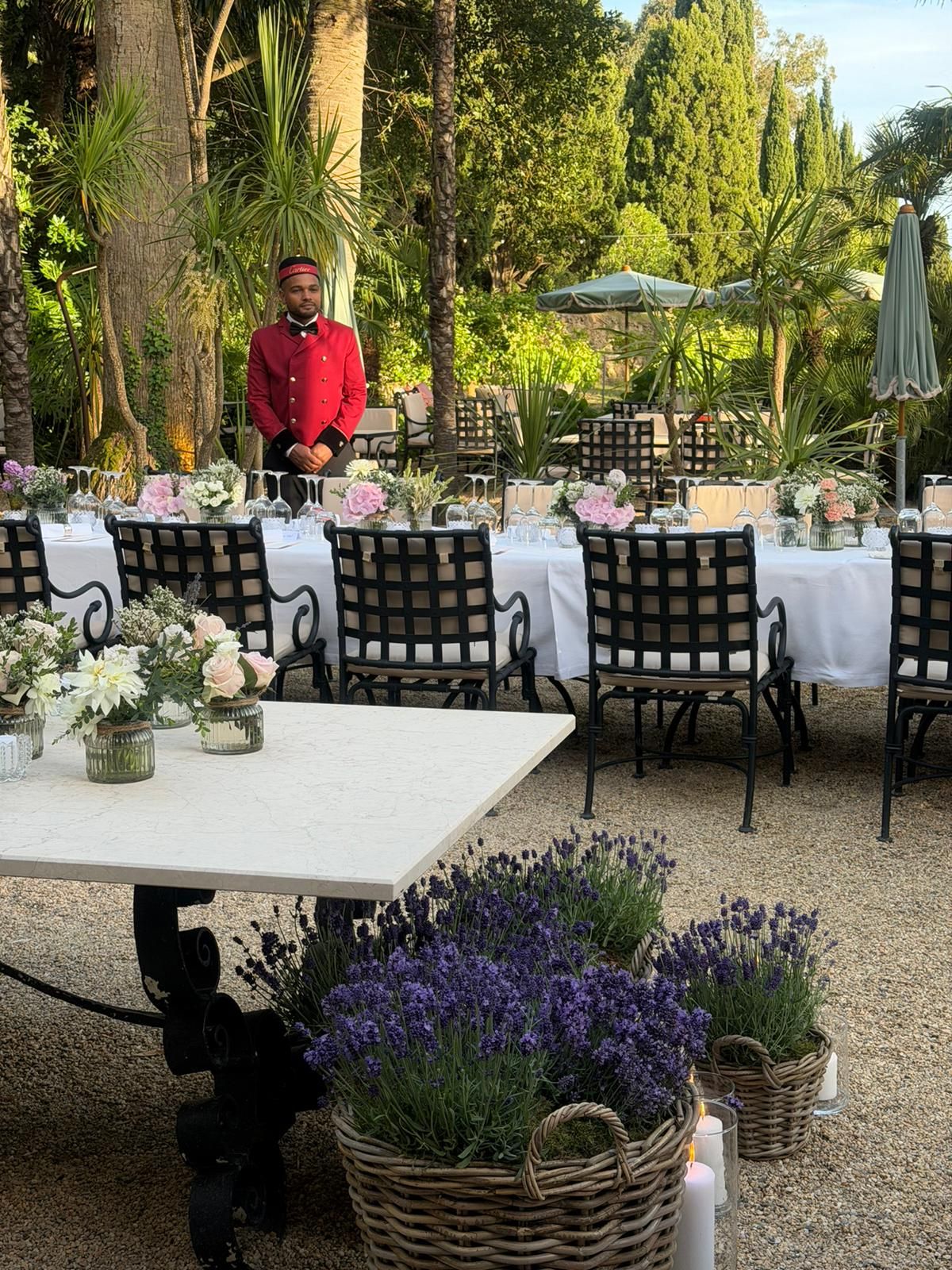 Outdoor garden dining setup with white tables, floral centerpieces, and a bellman in red uniform.