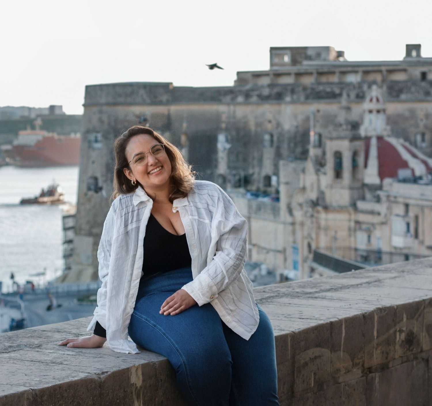 Smiling woman in white shirt and jeans sitting on a stone wall with historic Valletta Malta waterfront behind her.