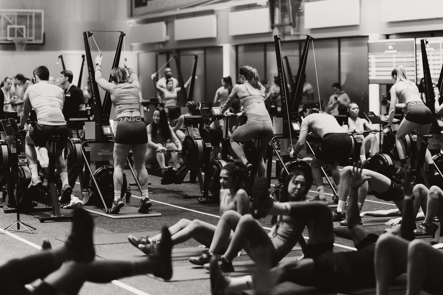 Black and white photo of a busy gym class with people exercising on stationary bikes and floor mats.