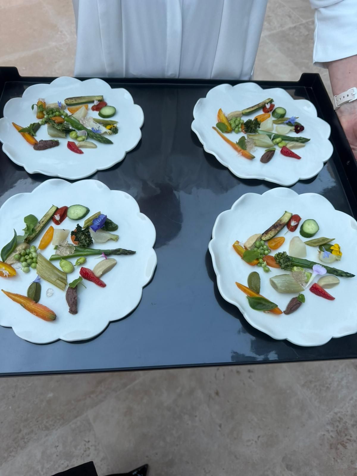 Four elegant white scalloped plates with colorful fresh vegetable and edible flower gourmet appetizers on serving tray.