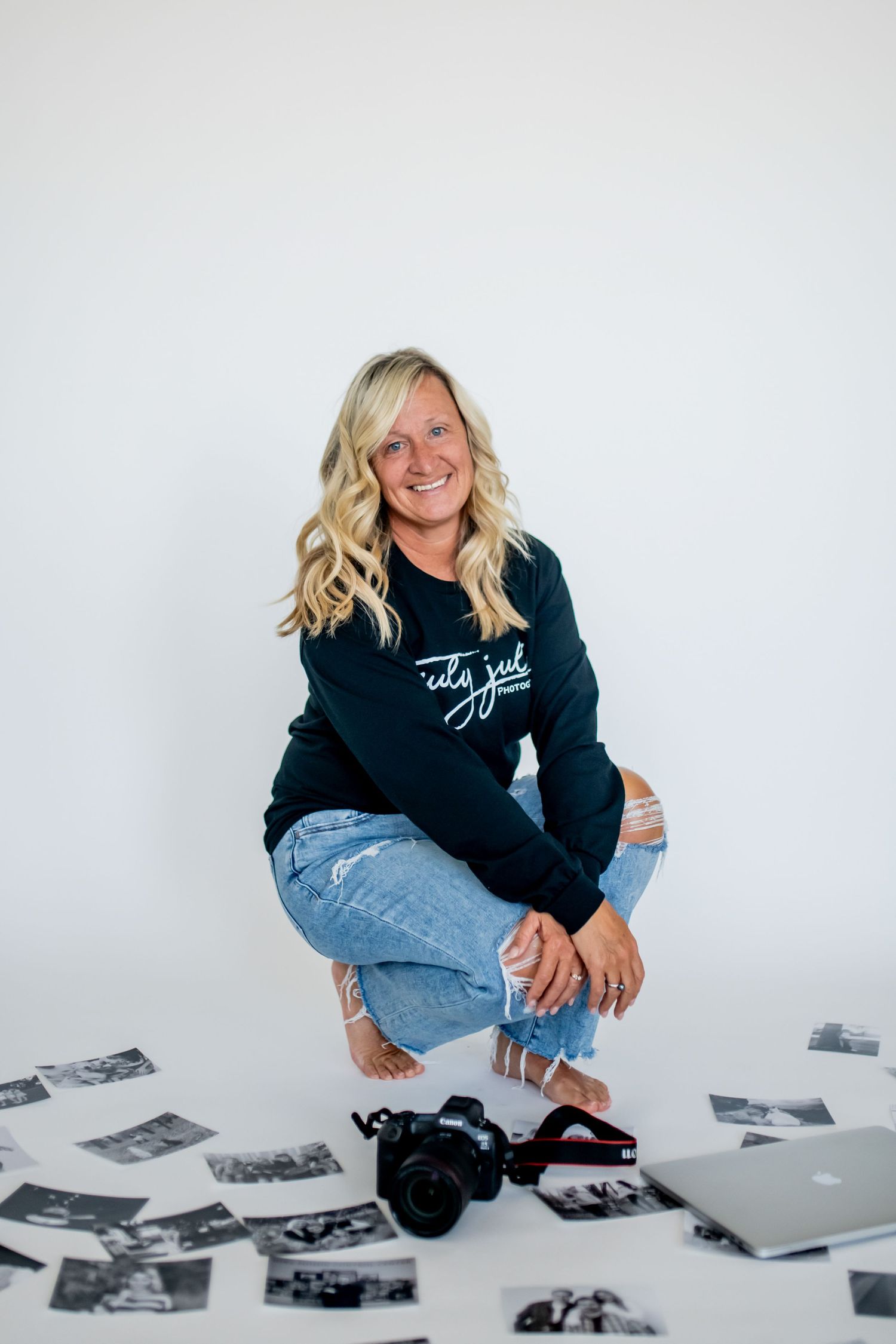 Photographer crouches on floor surrounded by scattered black and white photos, camera and laptop while wearing casual shirt and jeans.