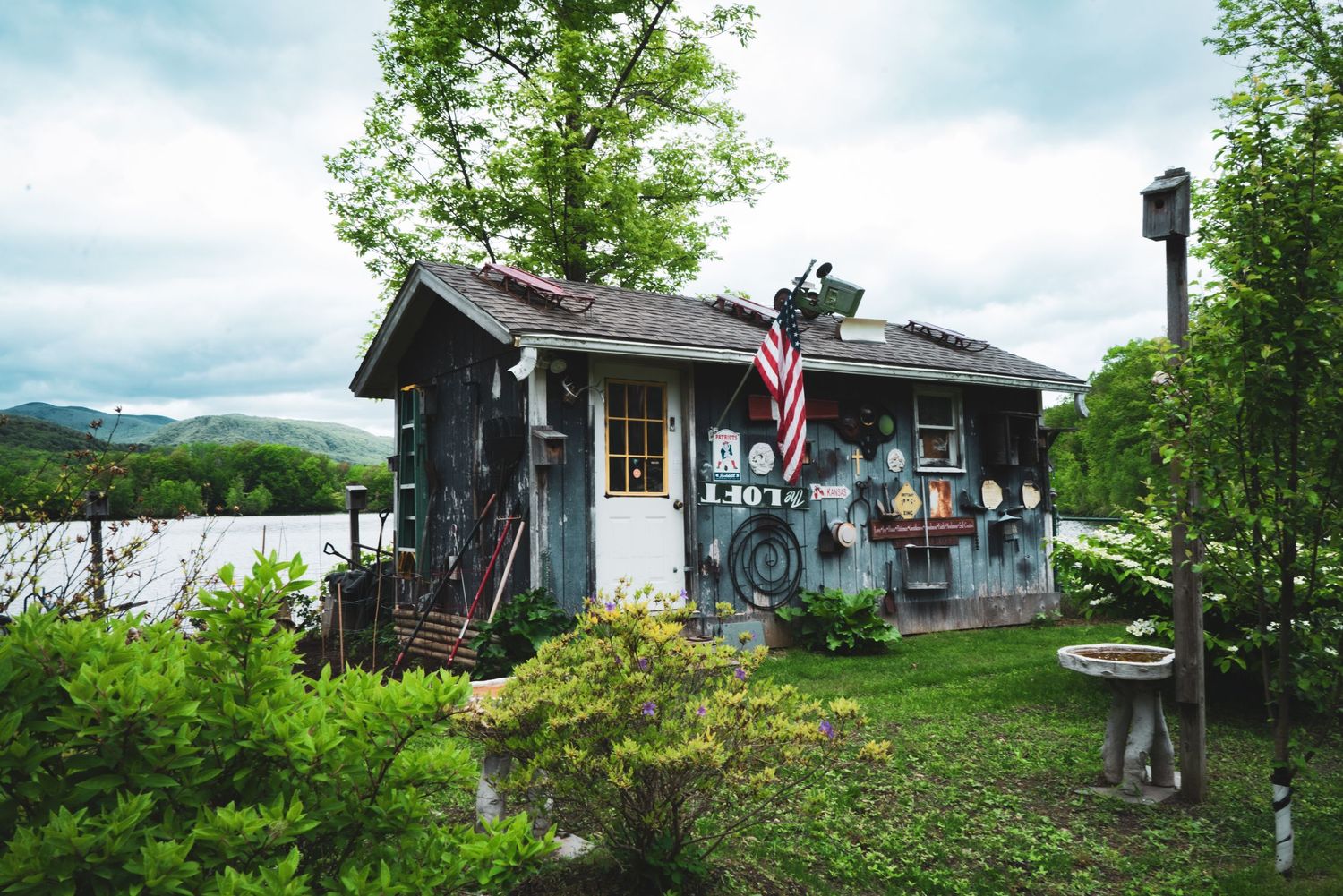 Charming rustic cottage with American flag, surrounded by lush green trees near a lake.