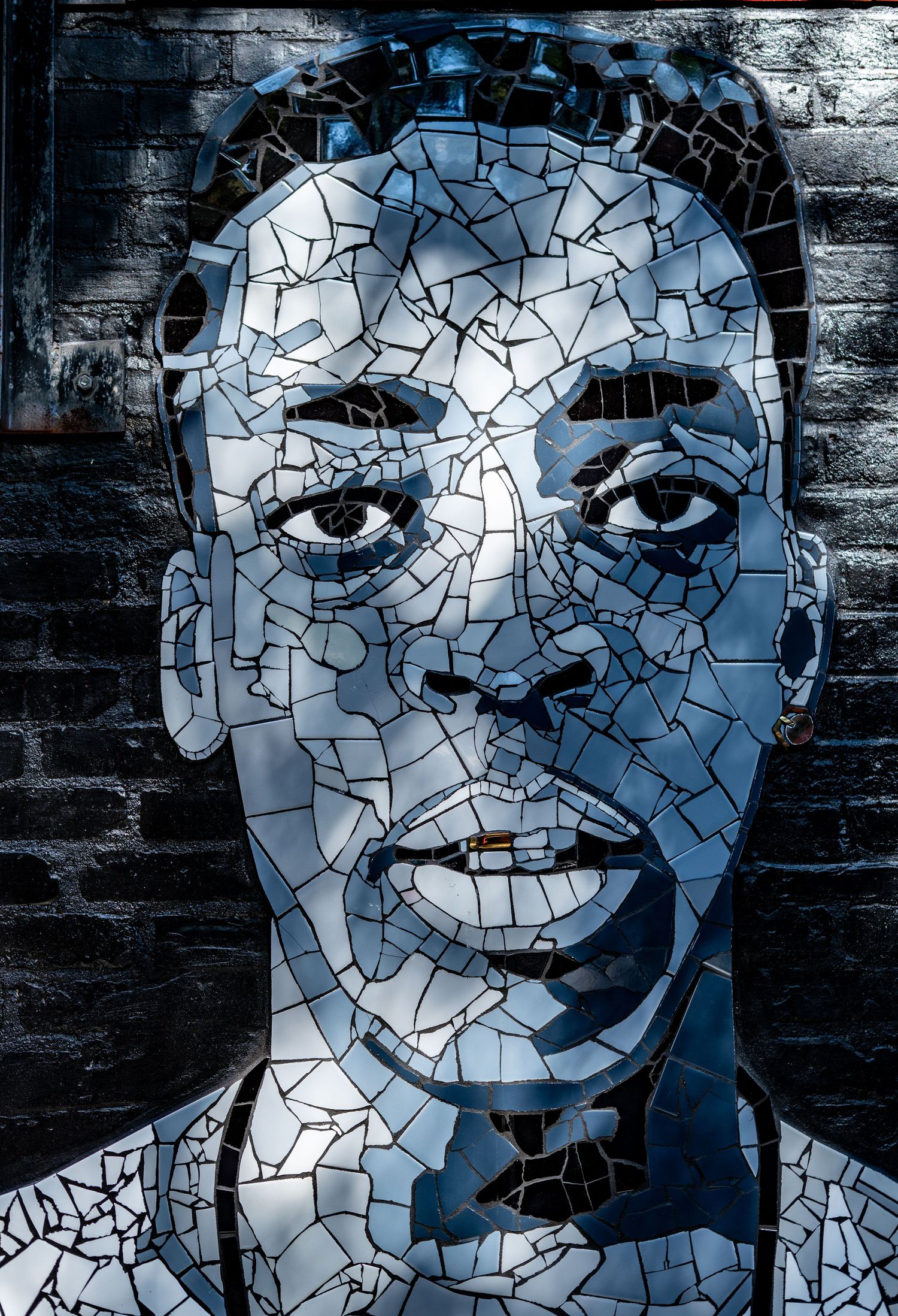 Mosaic portrait of a man made from broken mirror pieces mounted on a dark brick wall.