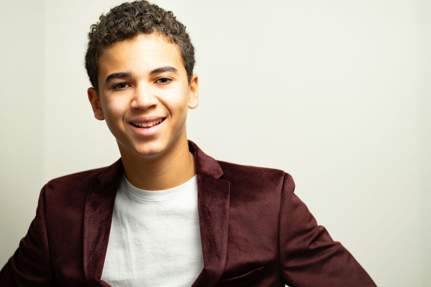 Smiling young man in a maroon blazer and white t-shirt posing against a light gray background.