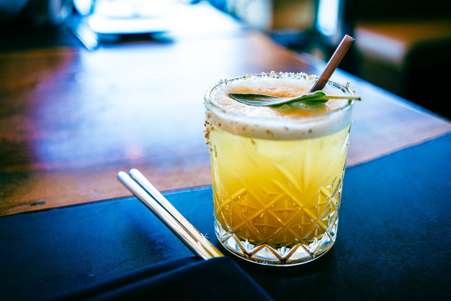 A crystal glass cocktail with orange juice, mint garnish, and a straw on a wooden bar.