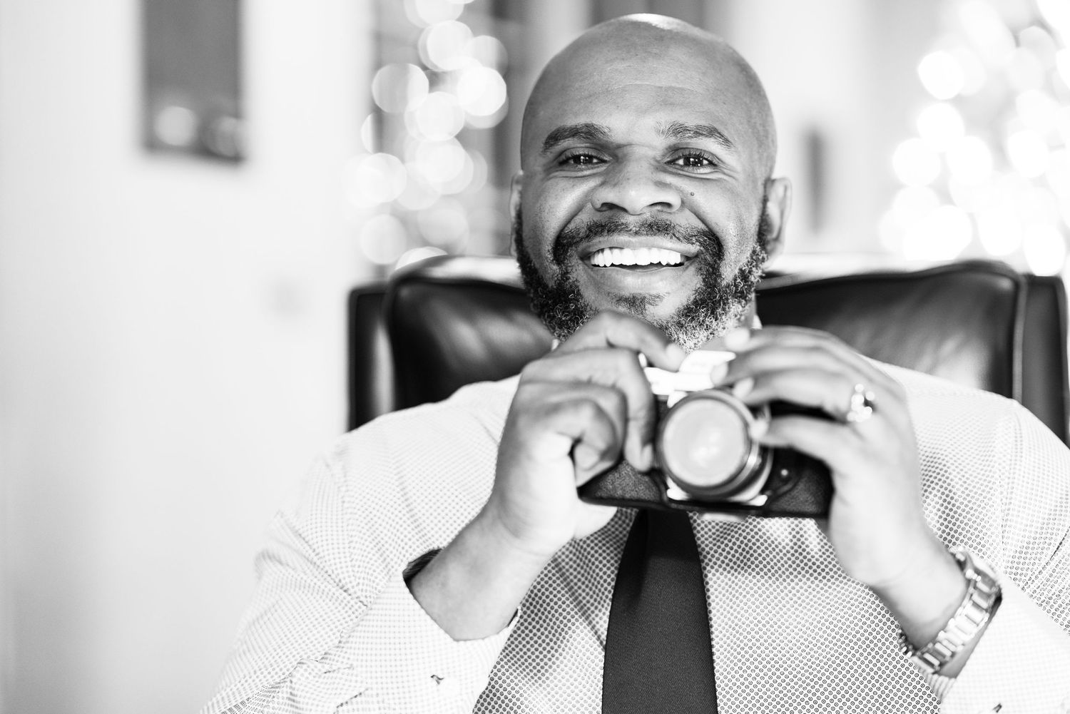 Black and white portrait of a smiling bald man holding a camera while sitting in a chair.