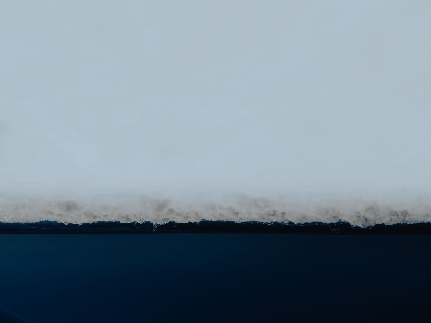 Minimalist landscape showing a thin layer of snow on ice with dark water below pale sky.