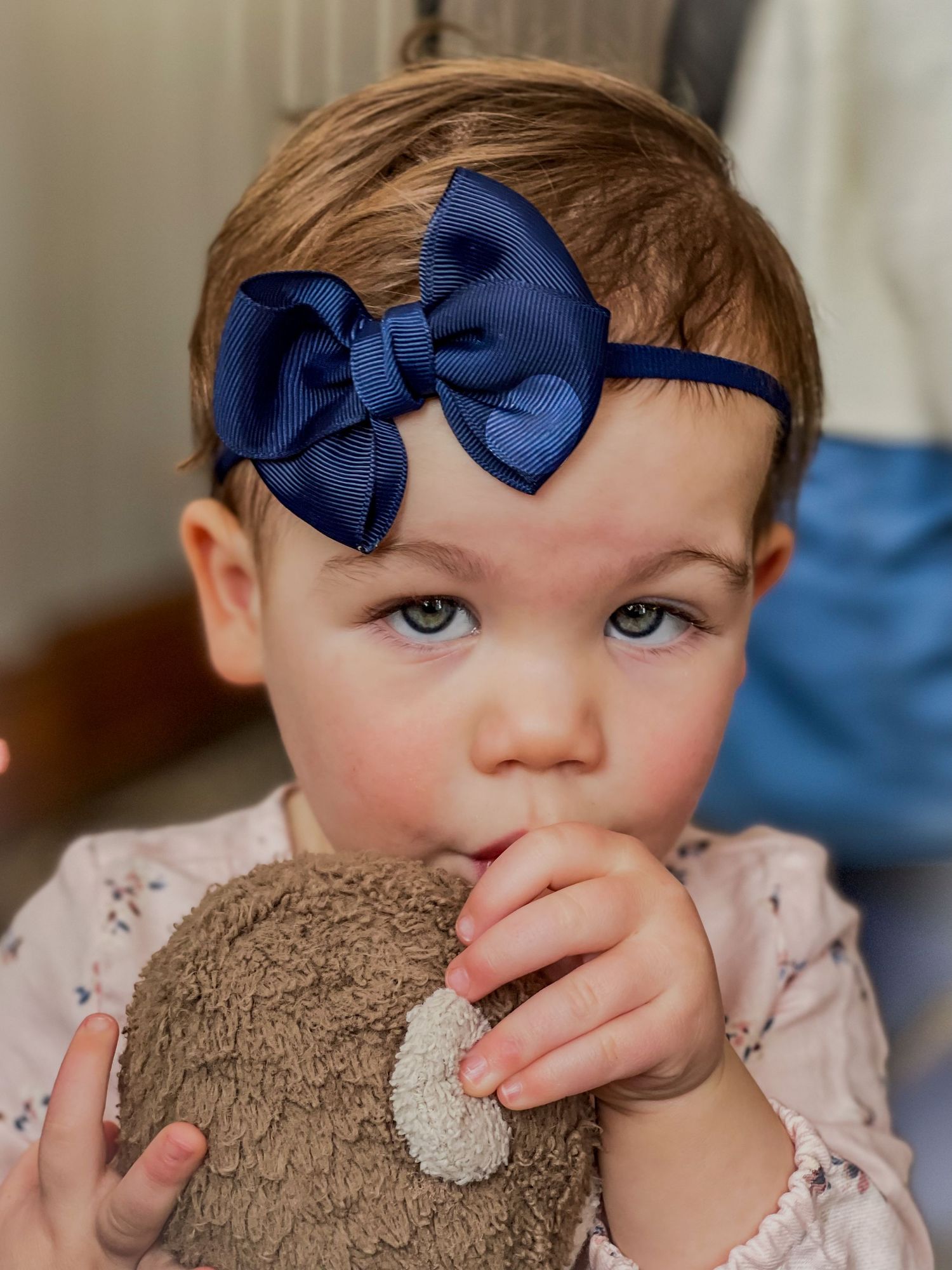 Adorable baby girl wearing a navy blue bow headband while chewing on a stuffed teddy bear.
