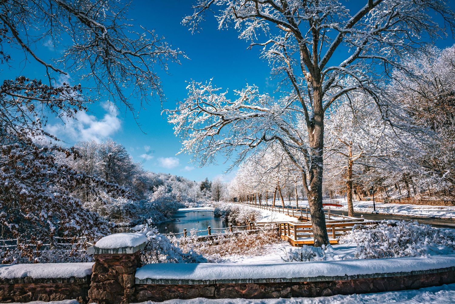 Snow-covered trees and stone fence surround a serene blue river under a bright winter sky.