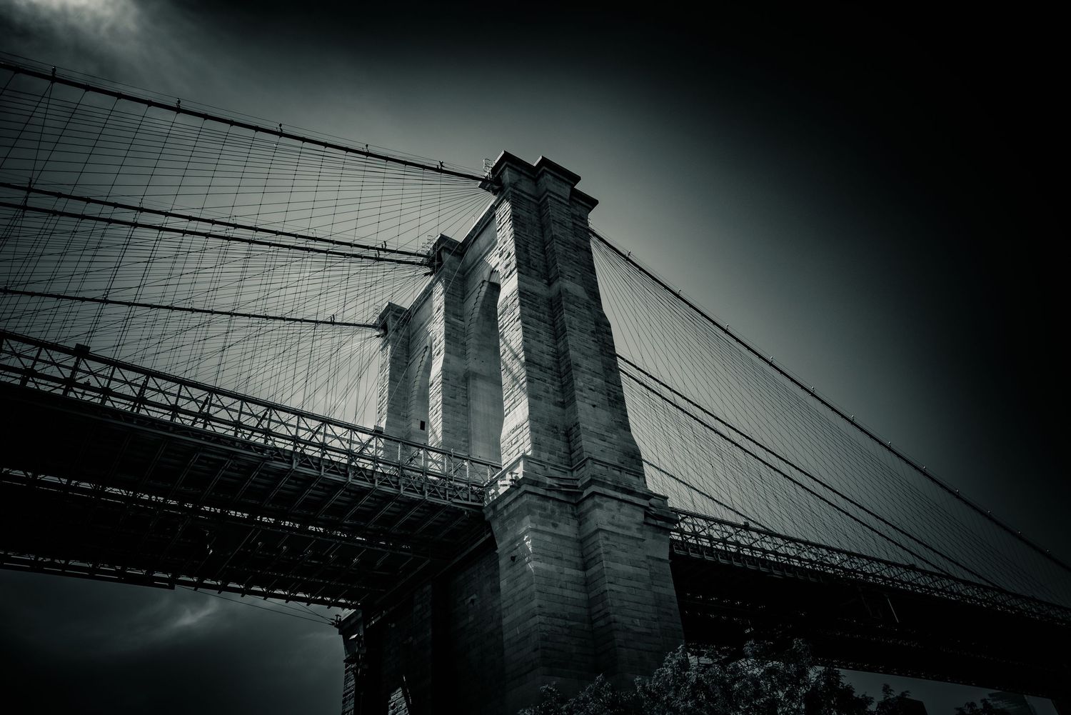 A dramatic low-angle black and white view of the Brooklyn Bridge towers against stormy sky.