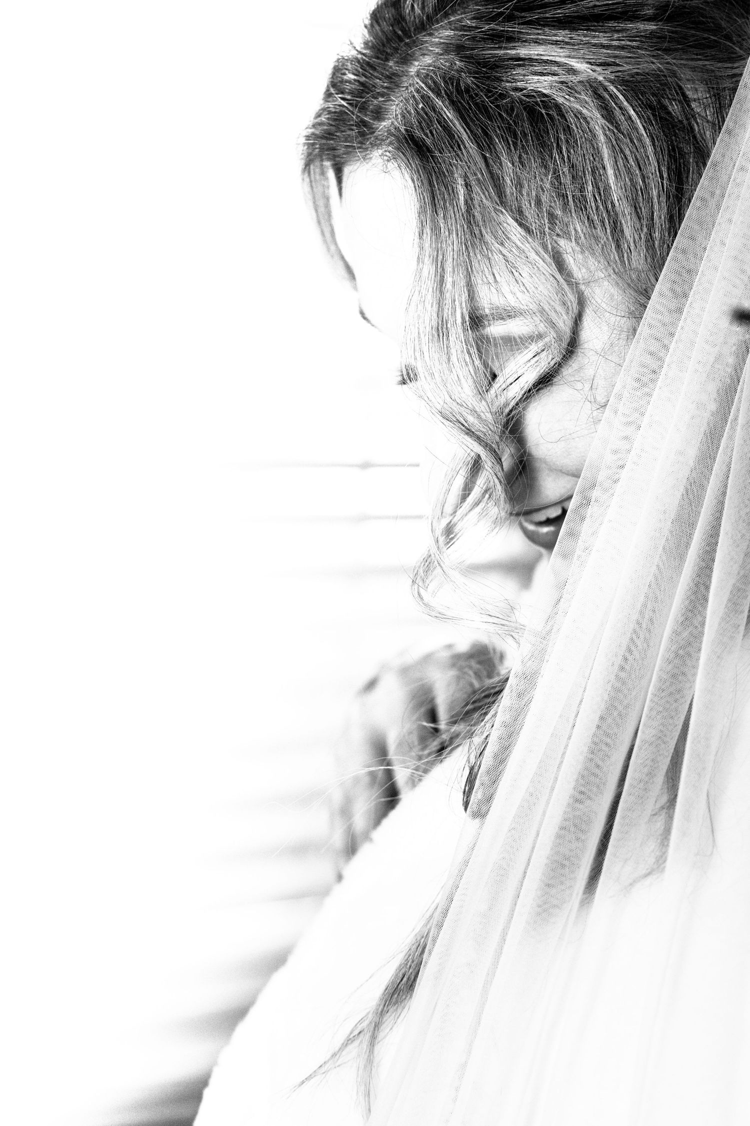 A black and white portrait of a bride with flowing veil and curly hair looking downward gracefully.