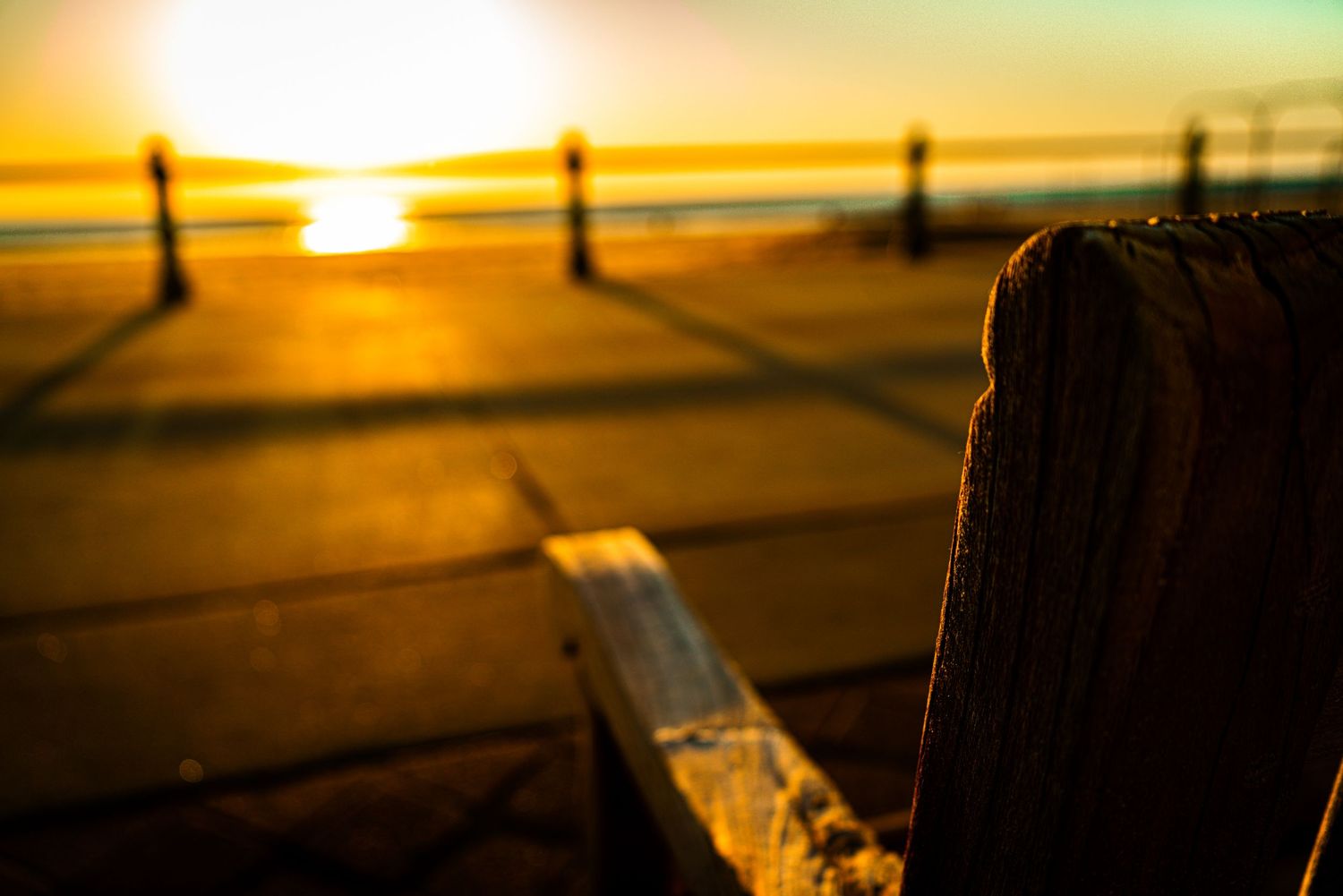 A wooden bench in the foreground with a stunning golden sunset over a beachside promenade at dusk.